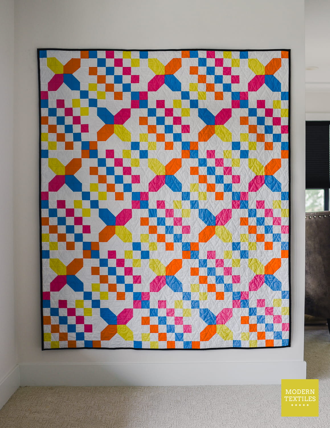 Checkered Crossing quilt hanging on white wall in background. Modern Textiles logo in bottom right corner.