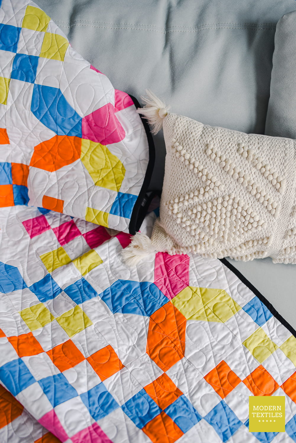 Checkered Crossing quilt draped over grey couch with off white woven throw pillow laid on top of quilt. Modern Textiles logo in bottom right corner.