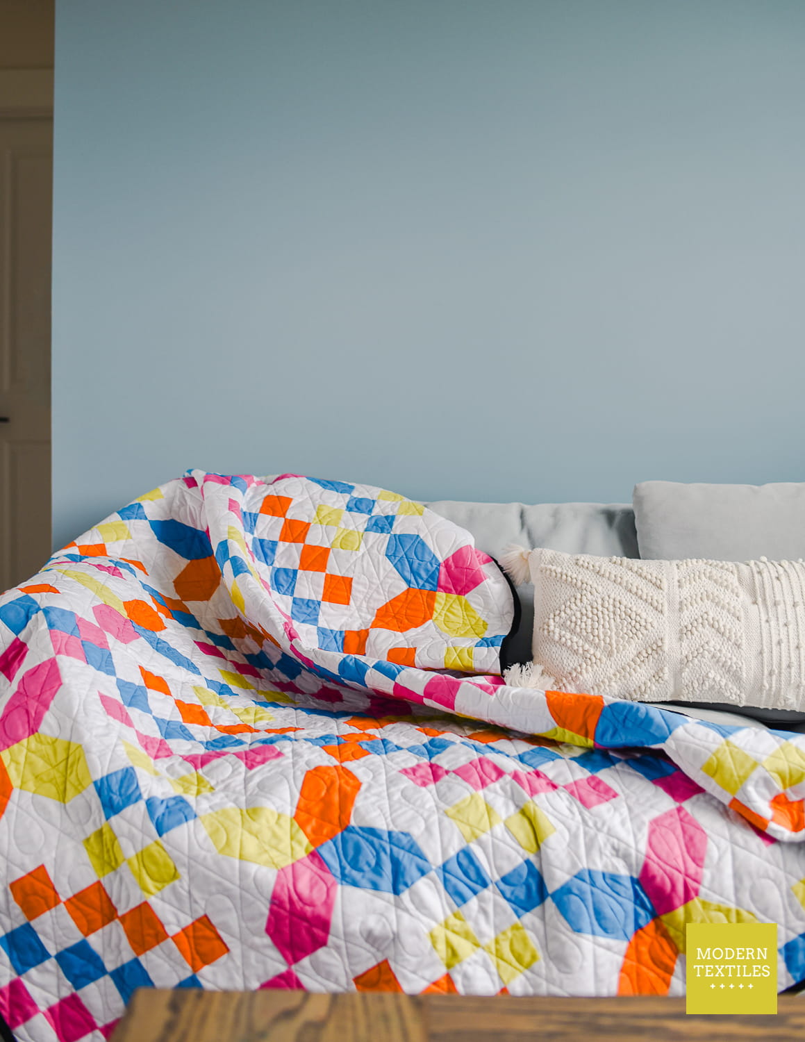 Checkered Crossing quilt drapped over a grey sofa, with a white texture throw picture on the right side of couch. Modern Textiles logo in bottom right corner.