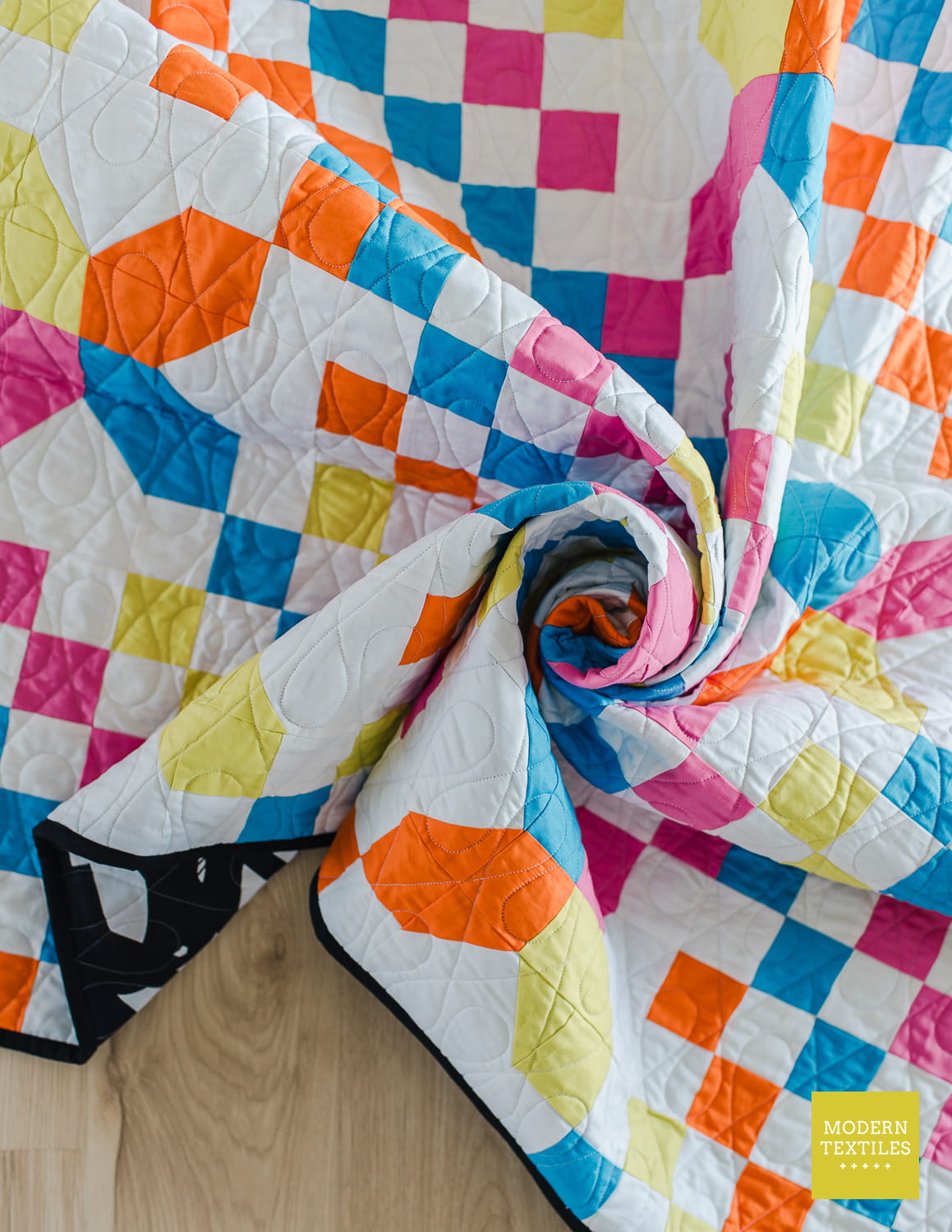 Checkered Crossing quilt laying on wood floor and swirled showing texture from quilting. Modern Textiles logo in bottom right corner.