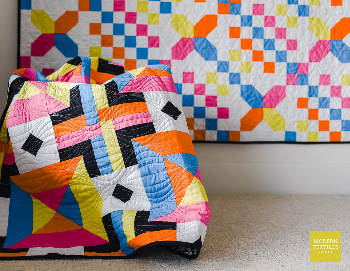 Checkered Crossing quilt hanging on white wall in background. Woven Squares quilt draped in basket in foreground. Modern Textiles logo in bottom right corner.