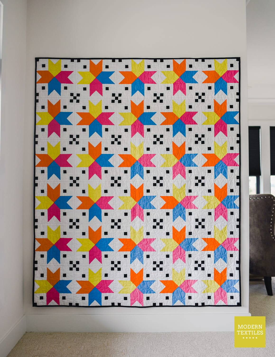 Four Corner Stars quilt hanging on white wall, with chair in background.