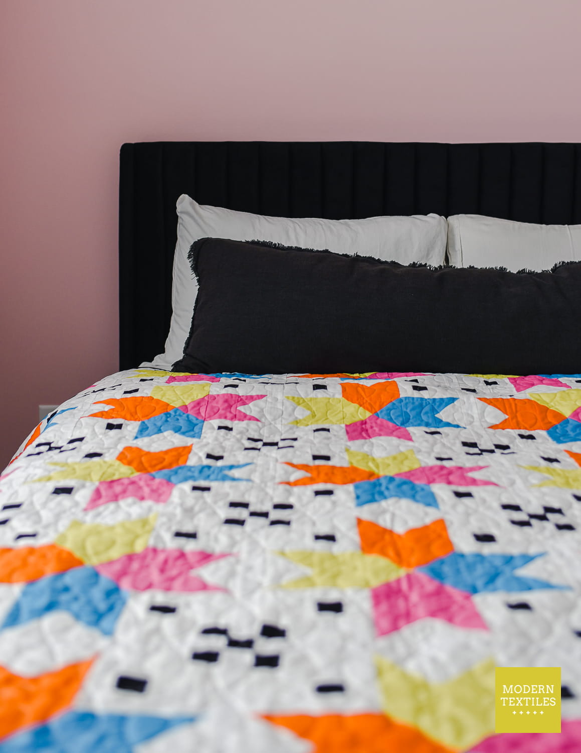 Colorful geometric quilt on a bed with a black headboard, white pillows and a black body pillow against a light pink wall.