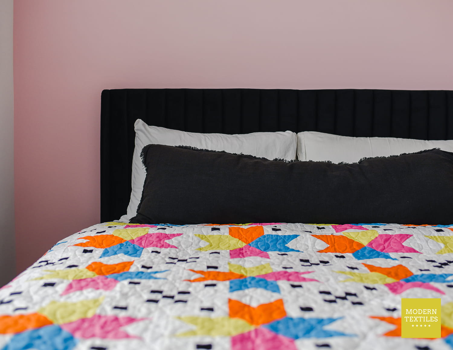 Colorful Four Corner Stars quilt with geometric pattern on a bed against a pink wall, with black and white pillows and black headboard featuring 'Modern Textiles' branding