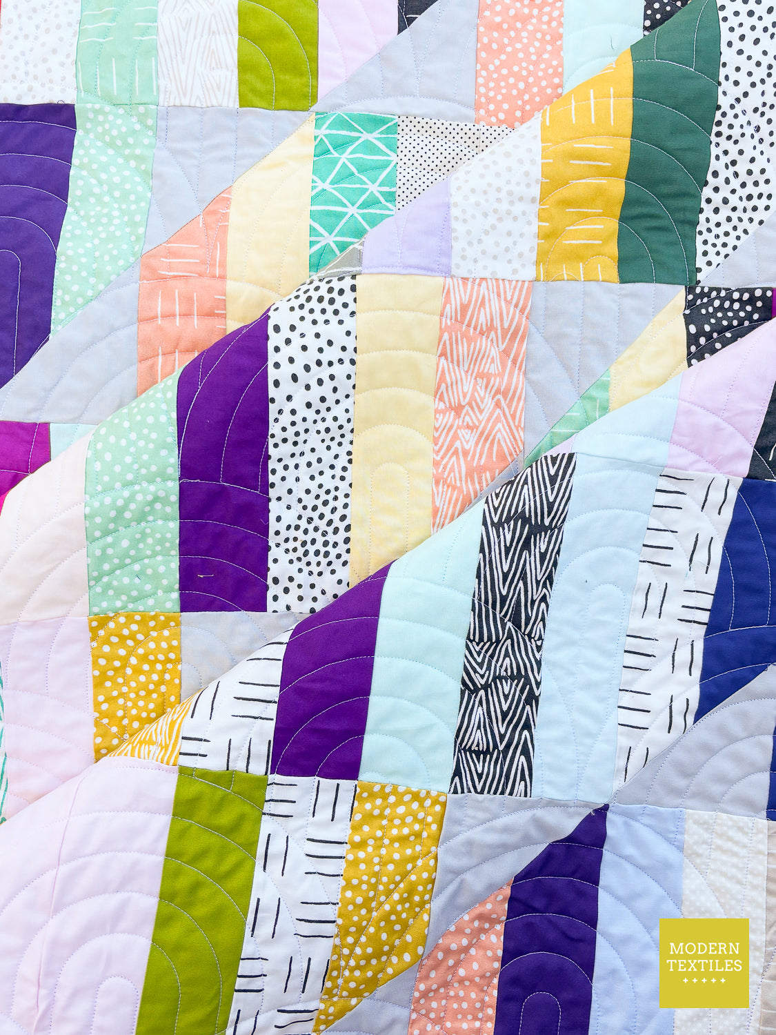A close-up of a colorful patchwork quilt with diagonal stitched sections of various fabrics in purple, green, yellow, pink, white, and black patterns. A small yellow 'MODERN TEXTILES' logo is visible in the bottom right corner.