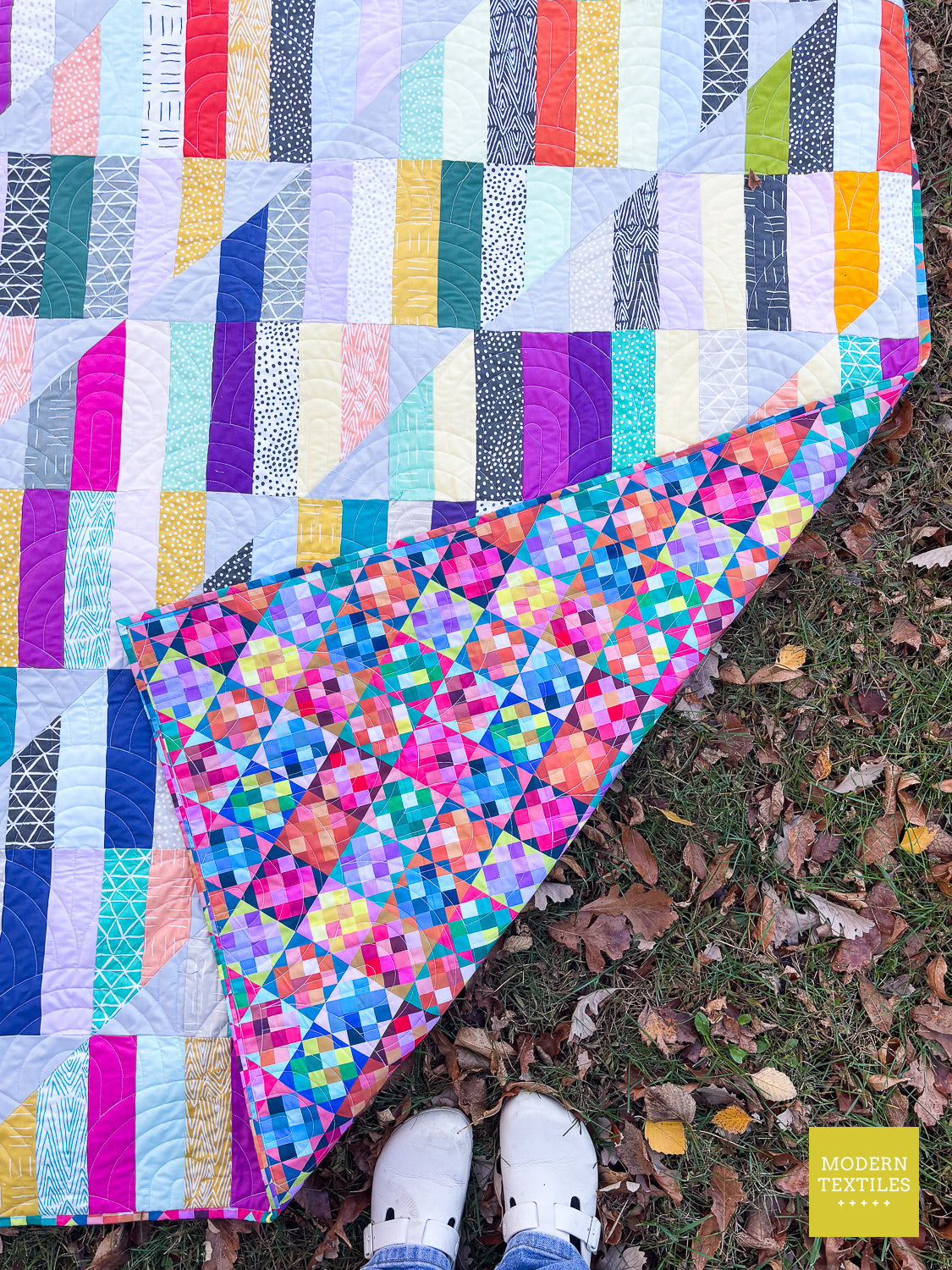 A colorful patchwork quilt laid out outdoors on grass with scattered leaves, with a person's white clogs visible at the bottom edge. The quilt features multiple patterned fabric panels in pink, blue, yellow, green, purple, and white.