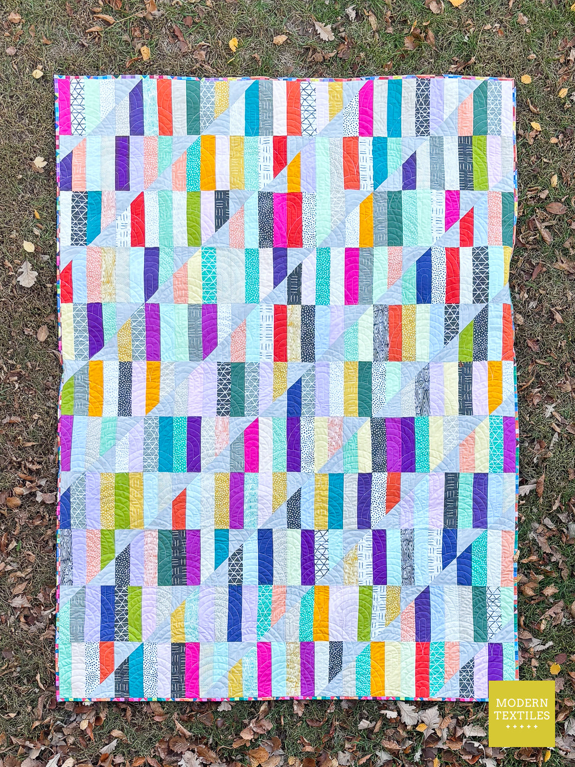 A colorful geometric patchwork quilt laid flat on grass with scattered autumn leaves. The quilt features a grid of vertical and diagonal striped blocks in a variety of bright colors including pink, blue, purple, green, yellow, orange, and white.