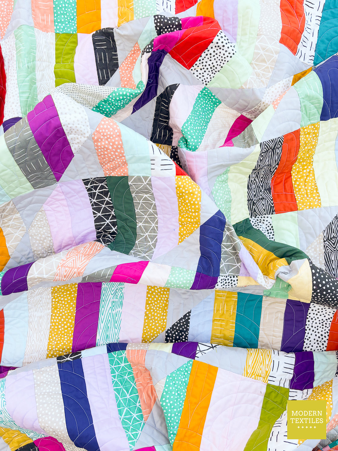 A close-up of a colorful patchwork quilt with geometric and striped fabric panels in purple, orange, green, pink, yellow, blue, white, and black patterns, with a small 'Modern Textiles' logo in the bottom right corner.