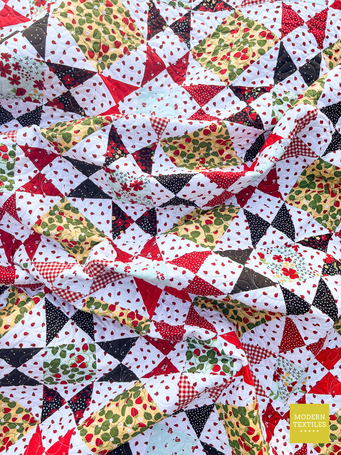 A close-up of a crinkled quilt showcasing Ohio Star Blocks made from fat quarter fabrics. Fabric design of red, black, green, and white triangles with various patterns including gingham, polka dots, and strawberry prints. Modern Textiles logo in lower right hand corner.