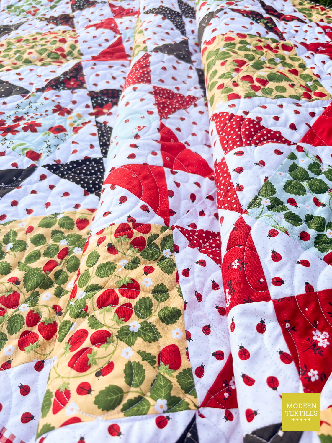 A close-up of a quilt featuring Ohio Star Blocks using strawberry and ladybug prints in red, green, yellow, and white. MODERN TEXTILES brand label is visible in the bottom right corner.