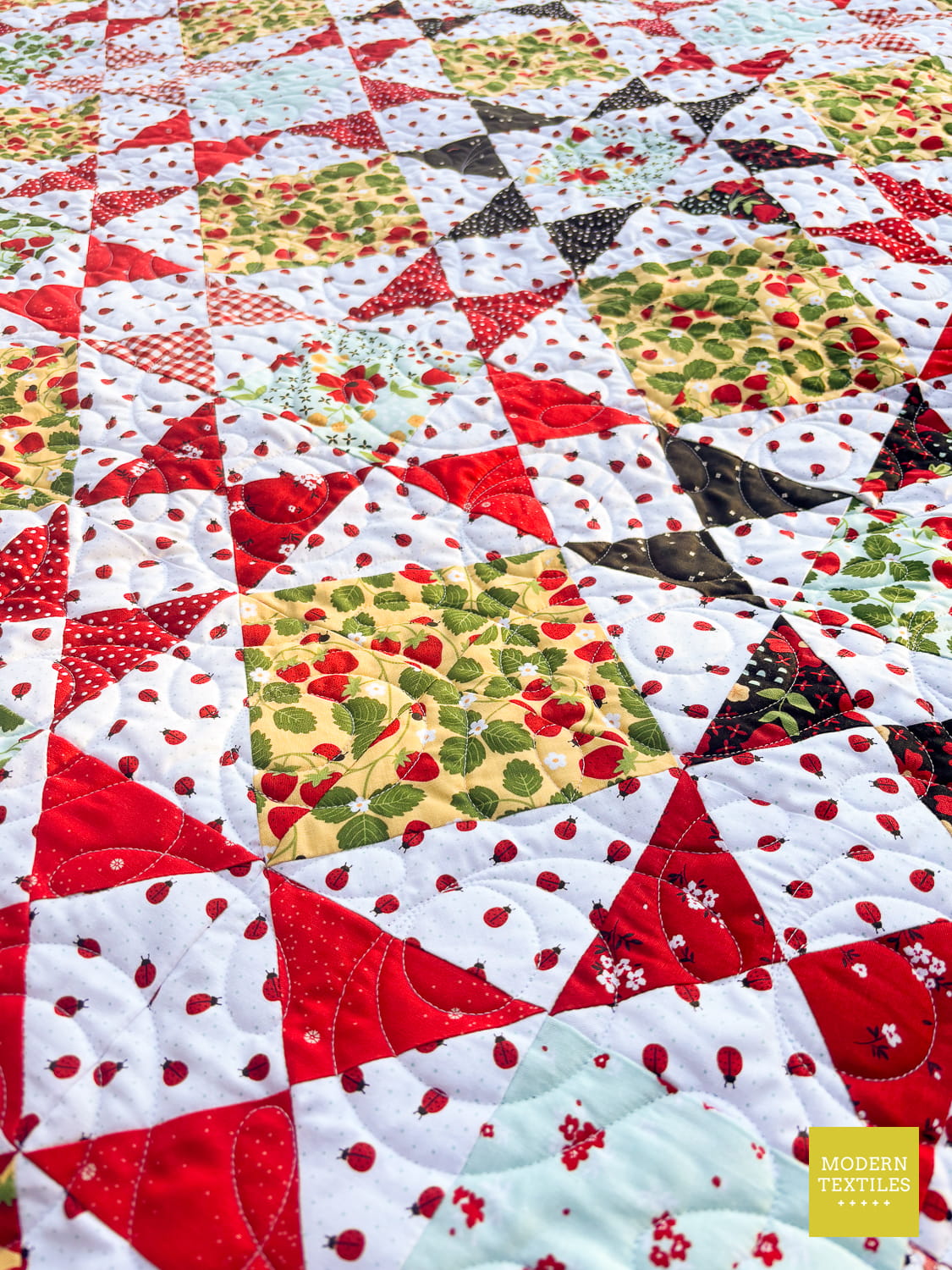 A close-up of a colorful quilt featuring Ohio Star Blocks and Half Square Triangle blocks using red strawberries on yellow backgrounds, red and black quarter square triangle blocks, and white fabric with small ladybugs. 'MODERN TEXTILES' logo is visible in the bottom right corner.