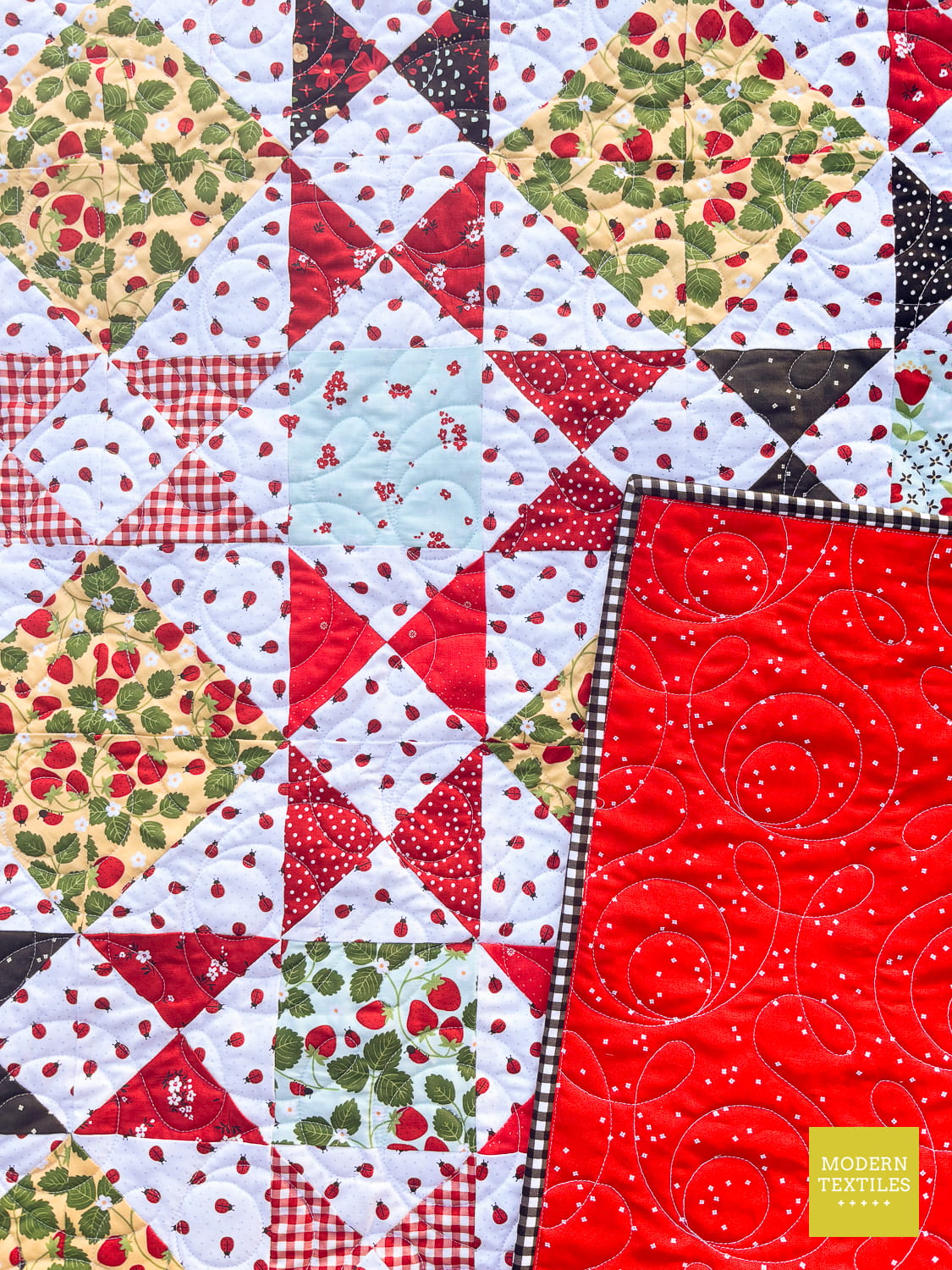 A close-up of the Ohio Star Exchange quilt fabric panel with a Ohio Star Block design featuring red, white, green, and black fabrics, plus a bright red quilt back with swirling quilting. A yellow Modern Textiles label is visible in the lower right corner.