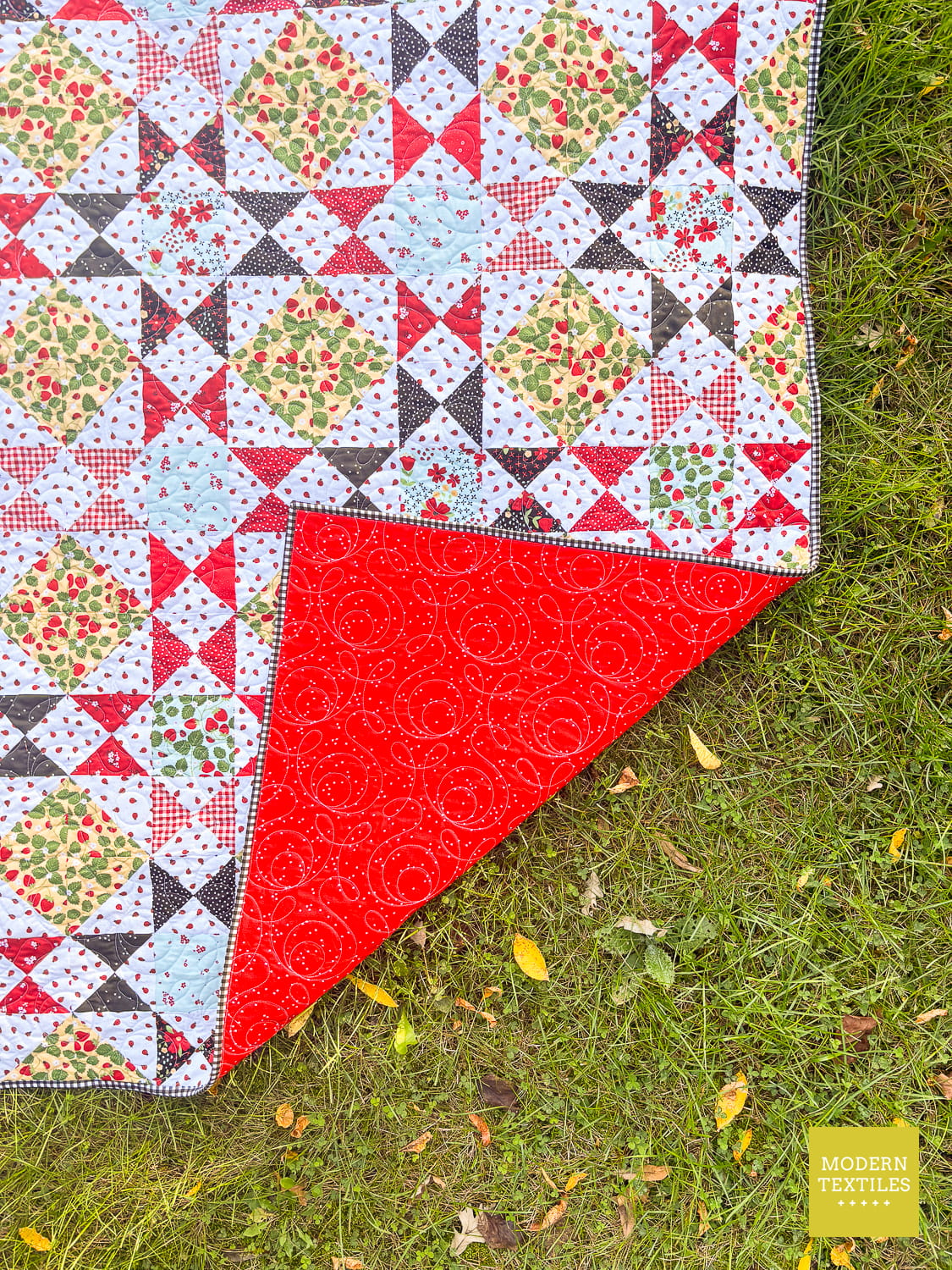 A quilt with colorful floral and geometric patchwork design is laid out on green grass. The blanket features a bright red reverse side with a Ginger Snap swirling pattern, and a corner is folded over to show the bright red quilt back fabric. Modern Textiles logo in lower right hand corner.