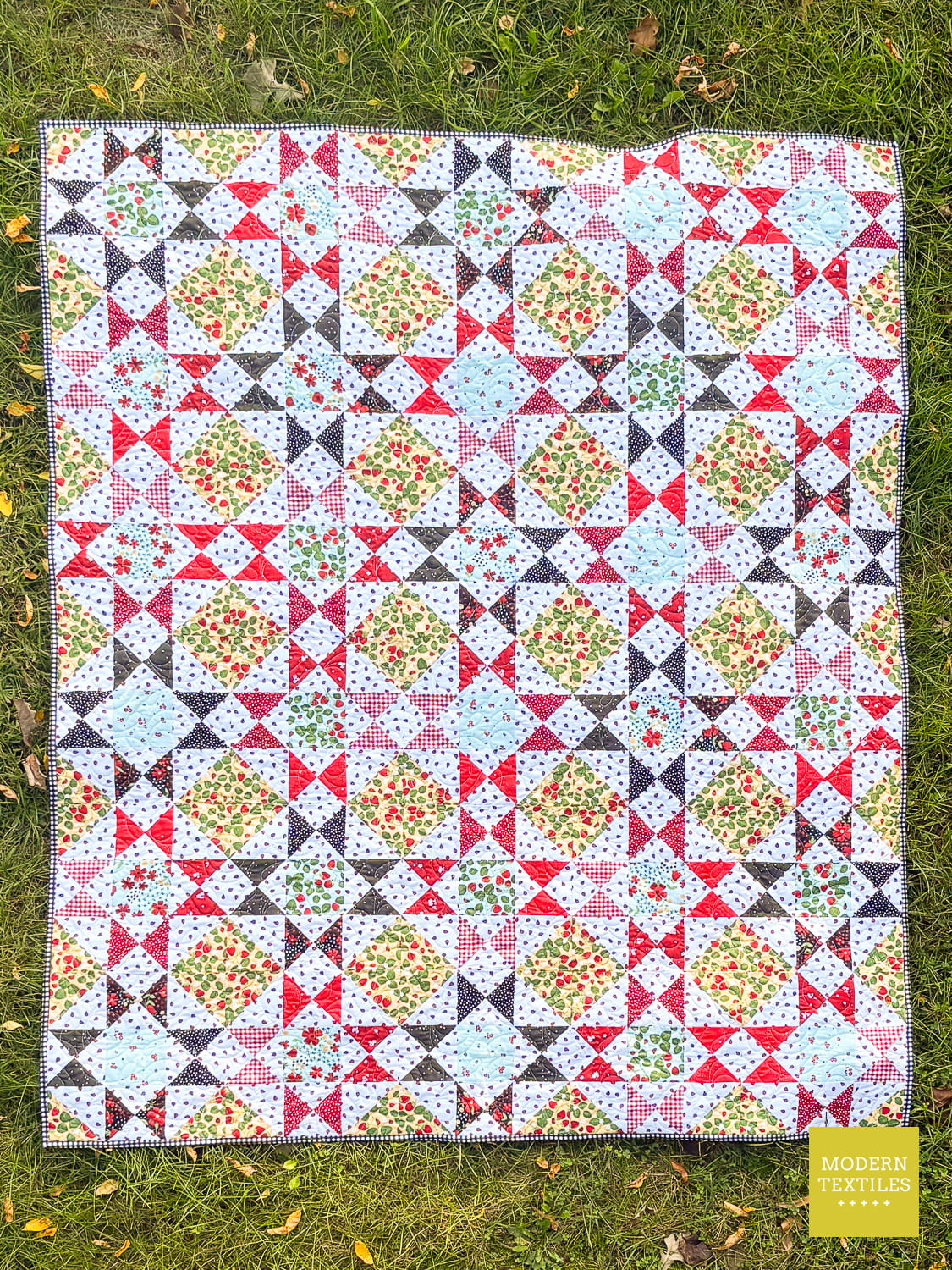 An Ohio Star Exchange quilt laid flat on green grass, featuring a repeating Ohio Star Block with Half Square Triangle patchwork pattern with red, black, light blue, yellow, printed fabrics. 'Modern Quiltiles' logo appears in the bottom right corner.