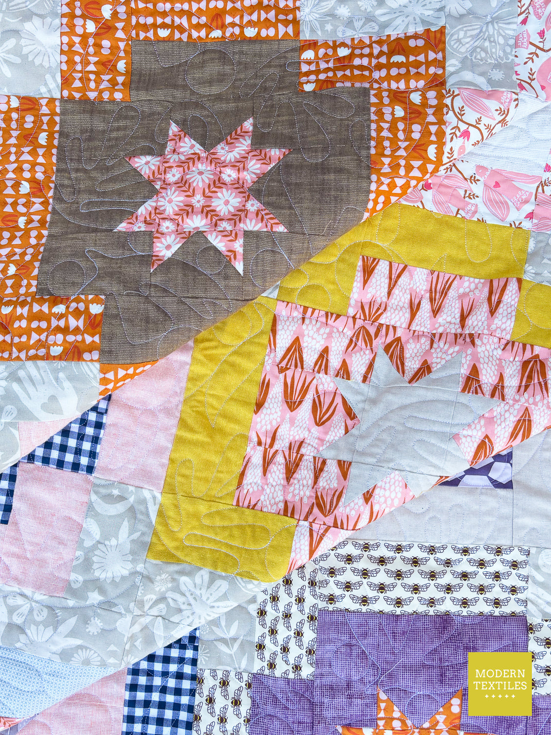A close-up of a colorful patchwork quilt with various fabric patterns including gingham, florals, stripes, and abstract prints in orange, pink, yellow, purple, brown, and white. A star-shaped patch is visible in the upper left, and a yellow block with orange abstract lines is in the lower right.