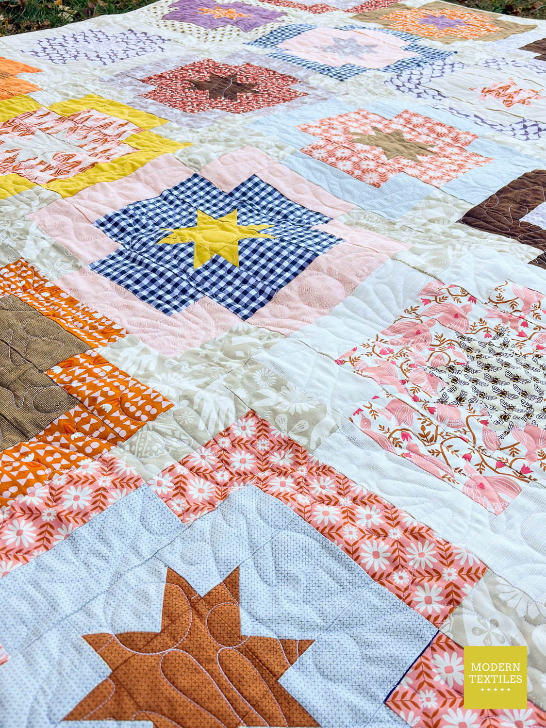 A colorful patchwork quilt spread out outdoors, featuring various fabric blocks, floral patterns, star and gingham designs, and quilted stitching. A yellow 'Modern Textiles' logo is visible in the bottom right corner.