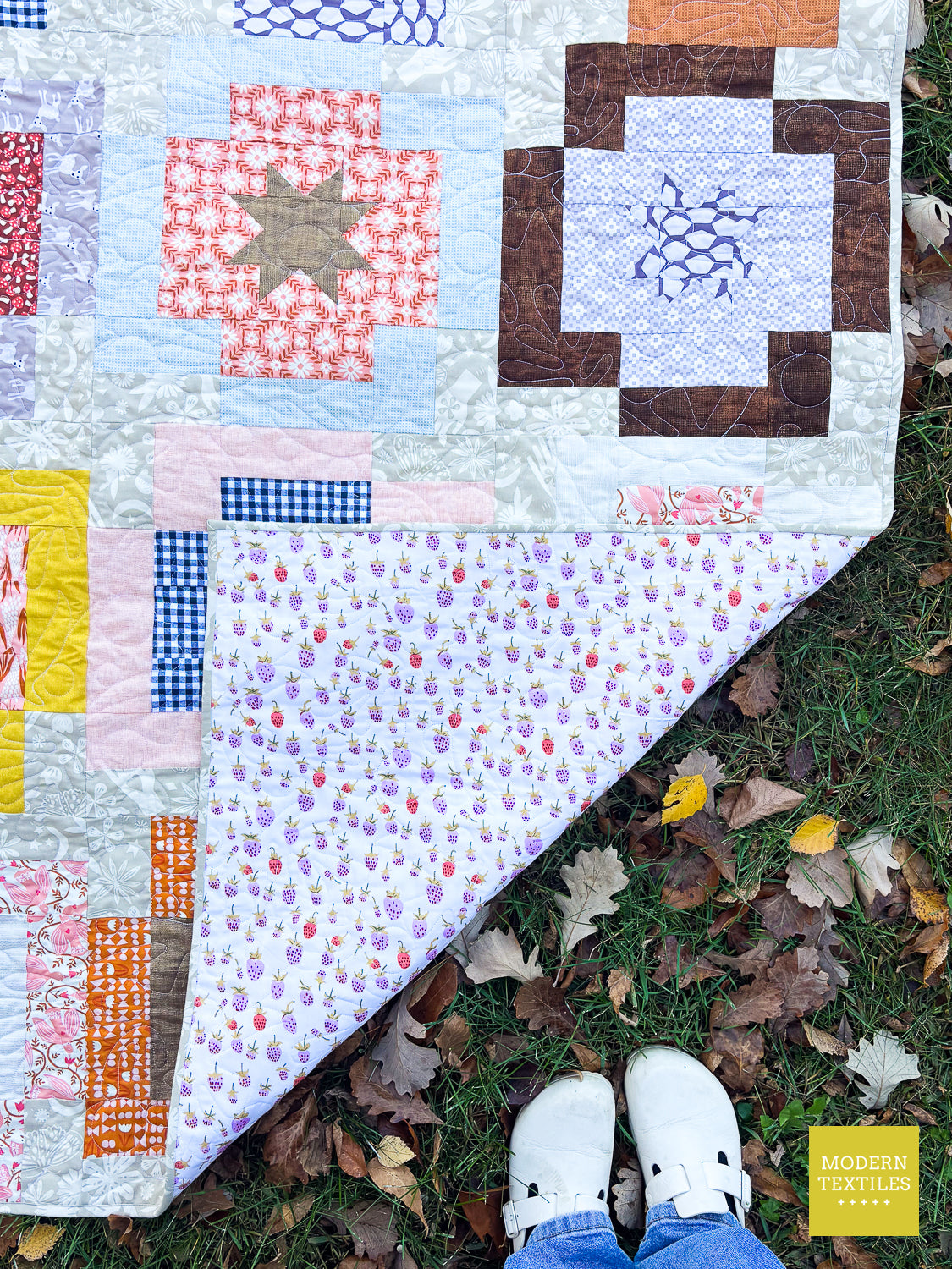 A multicolor patchwork quilt laid out on grass with fallen leaves, showing a person's white sneakers and blue jeans at the bottom edge. A yellow 'Modern Textiles' logo is visible in the bottom right corner.