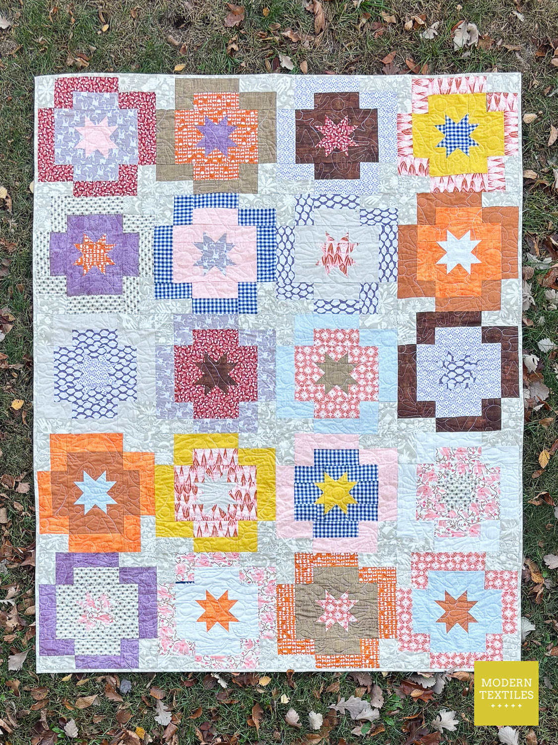 A colorful quilted patchwork blanket with various geometric and starburst block designs in multiple patterns, laid flat on a grassy lawn with fallen leaves. A yellow 'MODERN Textiles' logo is in the bottom right corner.