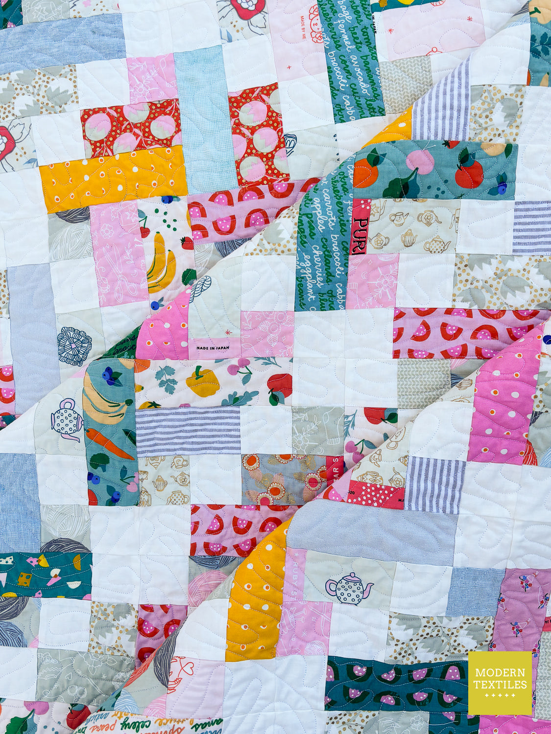 A close-up of a colorful patchwork quilt with various fabric patterns including fruit prints, stripes, polka dots, and floral designs. A yellow logo in the bottom right corner reads 'MODERN TEXTILES'.