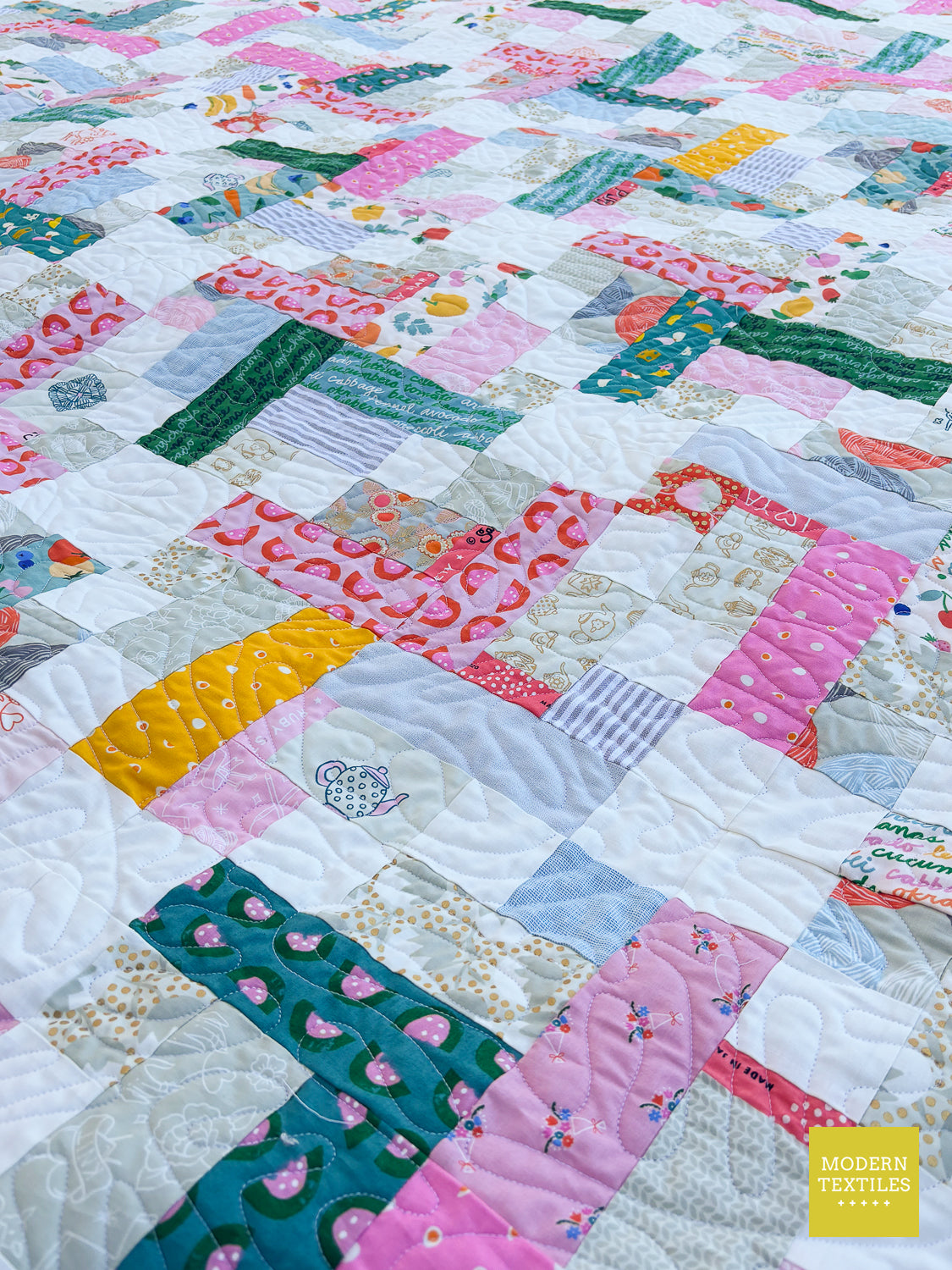 A close-up of a colorful patchwork quilt with various fabric blocks in pink, green, yellow, blue, and white, featuring a visible 'Modern Textiles' brand label in the bottom right corner.