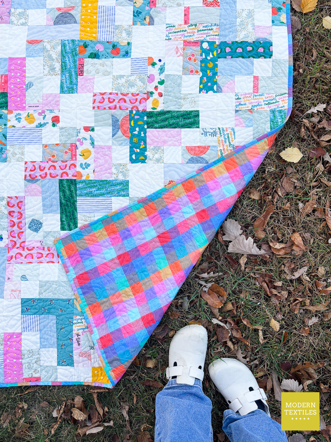 A colorful patchwork quilt laid on grass outdoors, with a person's legs and white shoes visible at the bottom. The quilt has a multicolor patchwork design on one side and a bright plaid pattern on the underside.