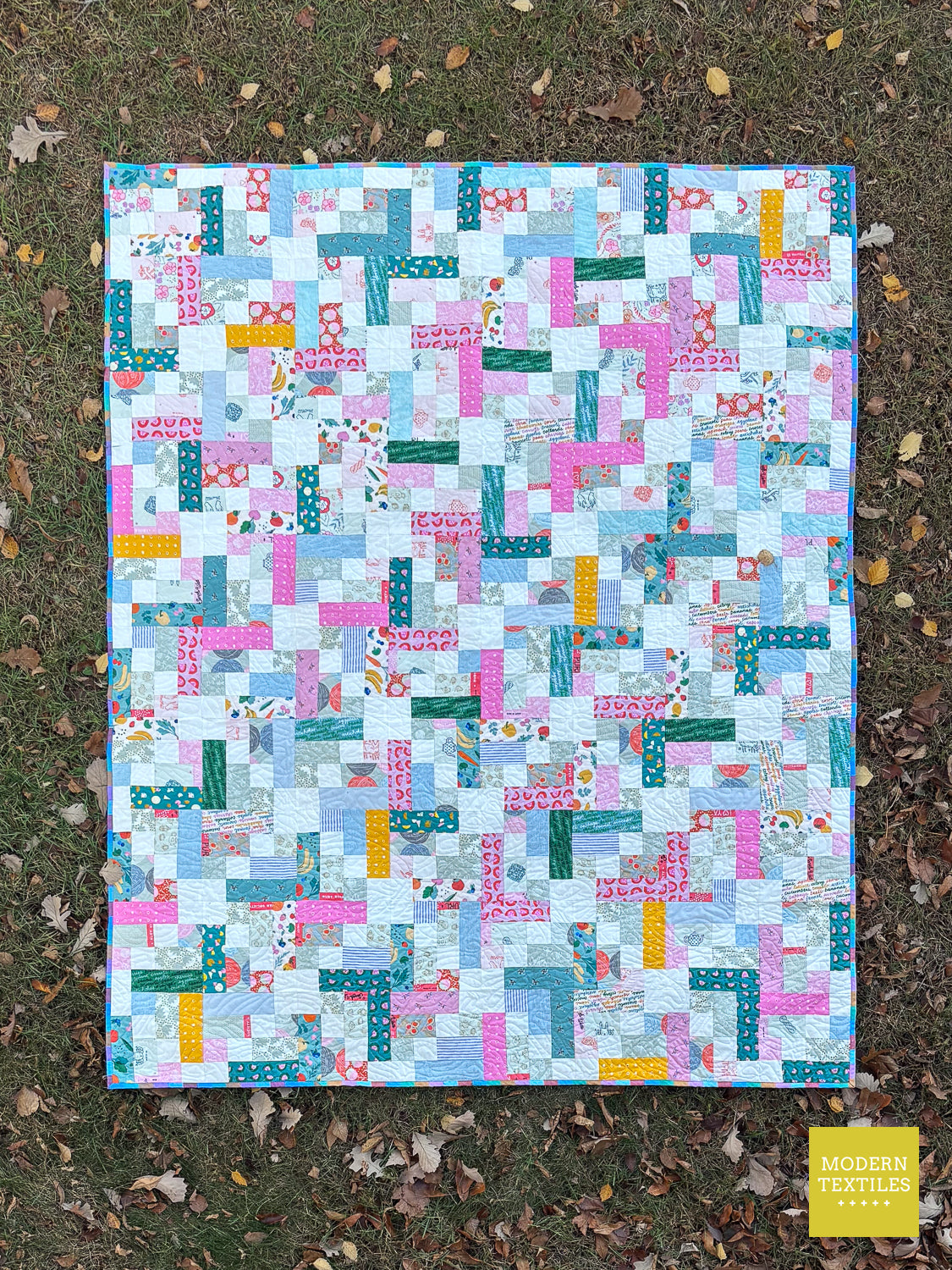 A colorful patchwork quilt laid flat on grass with scattered fallen leaves. The quilt features a geometric, block-like pattern in light blue, teal, pink, white, and small yellow accents.