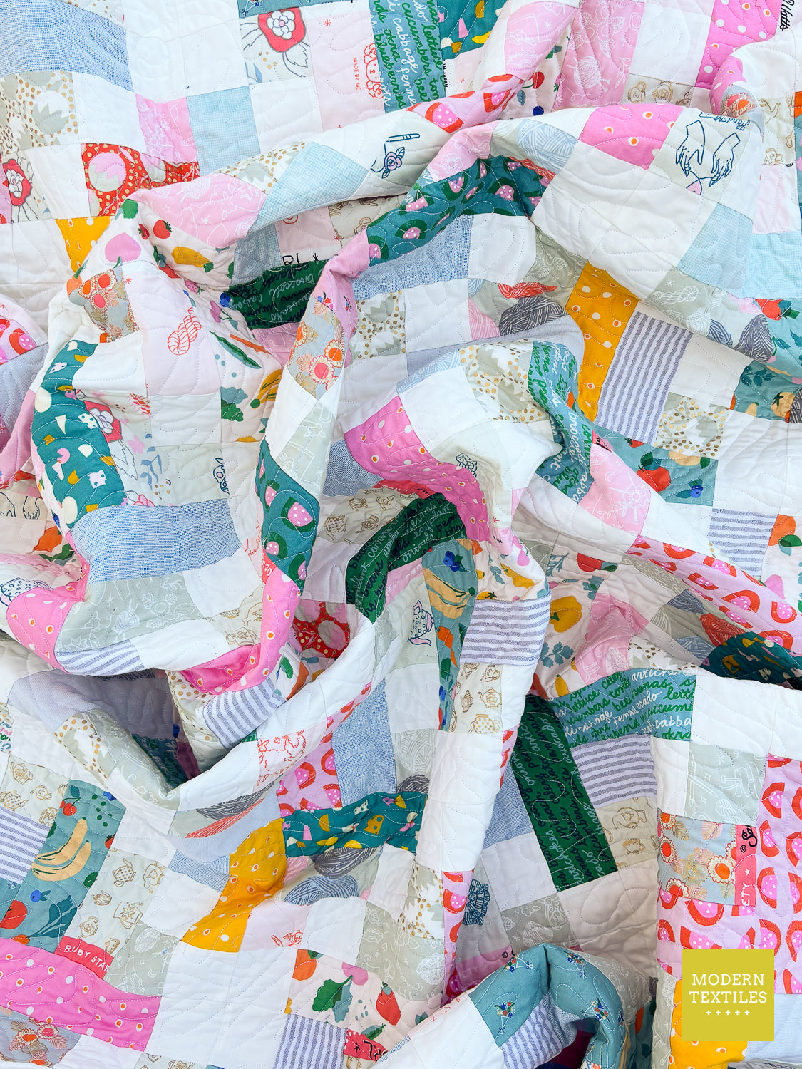 A close-up of a handmade patchwork quilt with a ruffled, gathered edge. The quilt features a variety of colorful, whimsical prints including strawberries, rainbows, animals, and patterns in pink, blue, green, yellow, and white.