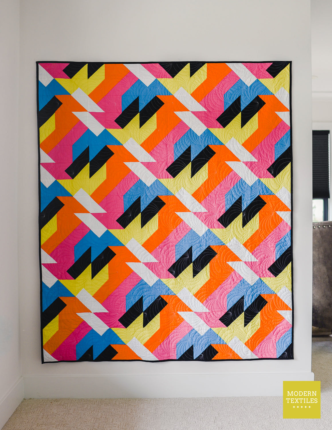 Colorful modern quilt pattern “Sheared” by Modern Textiles hanging on a wall, featuring bold geometric shapes in pink, orange, yellow, blue, black, and white.