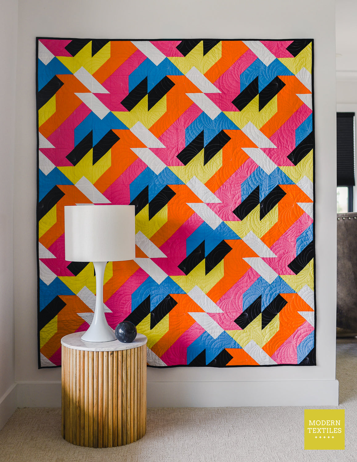 Modern quilt pattern “Sheared” displayed on a wall above a side table and lamp, highlighting colorful geometric patchwork in bright modern fabrics.