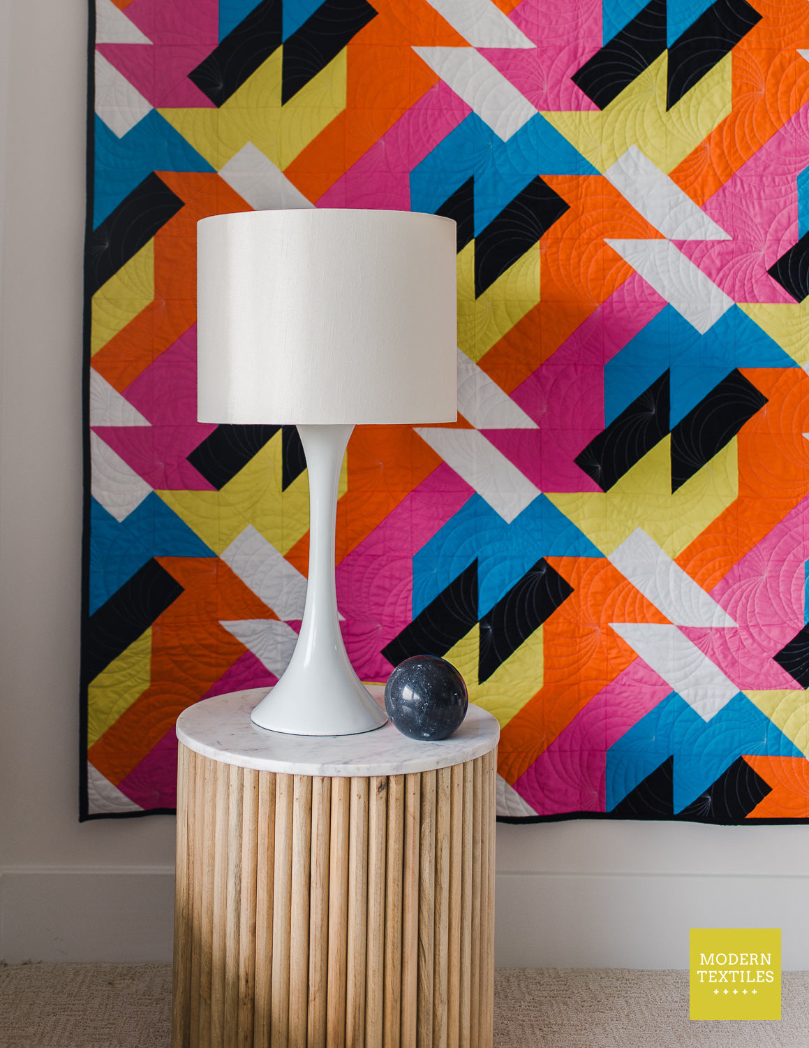 Modern geometric quilt “Sheared” styled in a living room with a lamp and round side table, featuring bright pink, orange, yellow, blue, and black fabrics.