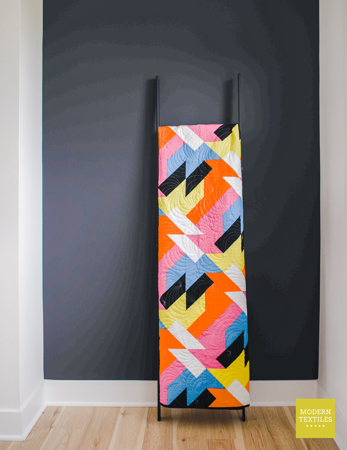 Tall vertical view of the Sheared modern quilt folded and hanging on a quilt ladder against a dark wall, showing bold geometric patchwork in vibrant colors.