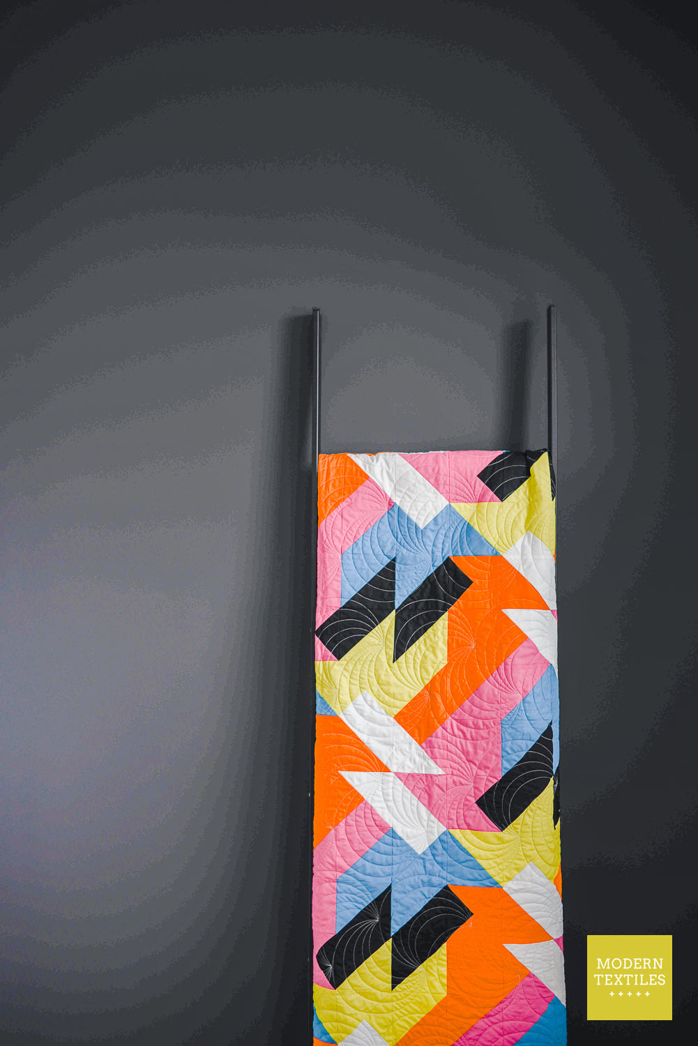 Modern quilt “Sheared” partially visible against a dark wall folded and hanging from a quilt ladder.