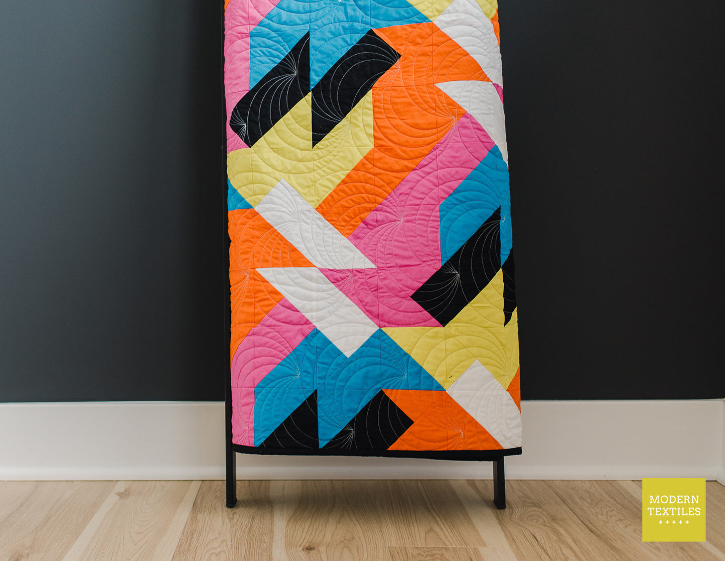 Sheared modern quilt draped over a quilt ladder, highlighting bold geometric blocks in bright pink, orange, yellow, blue, and black.