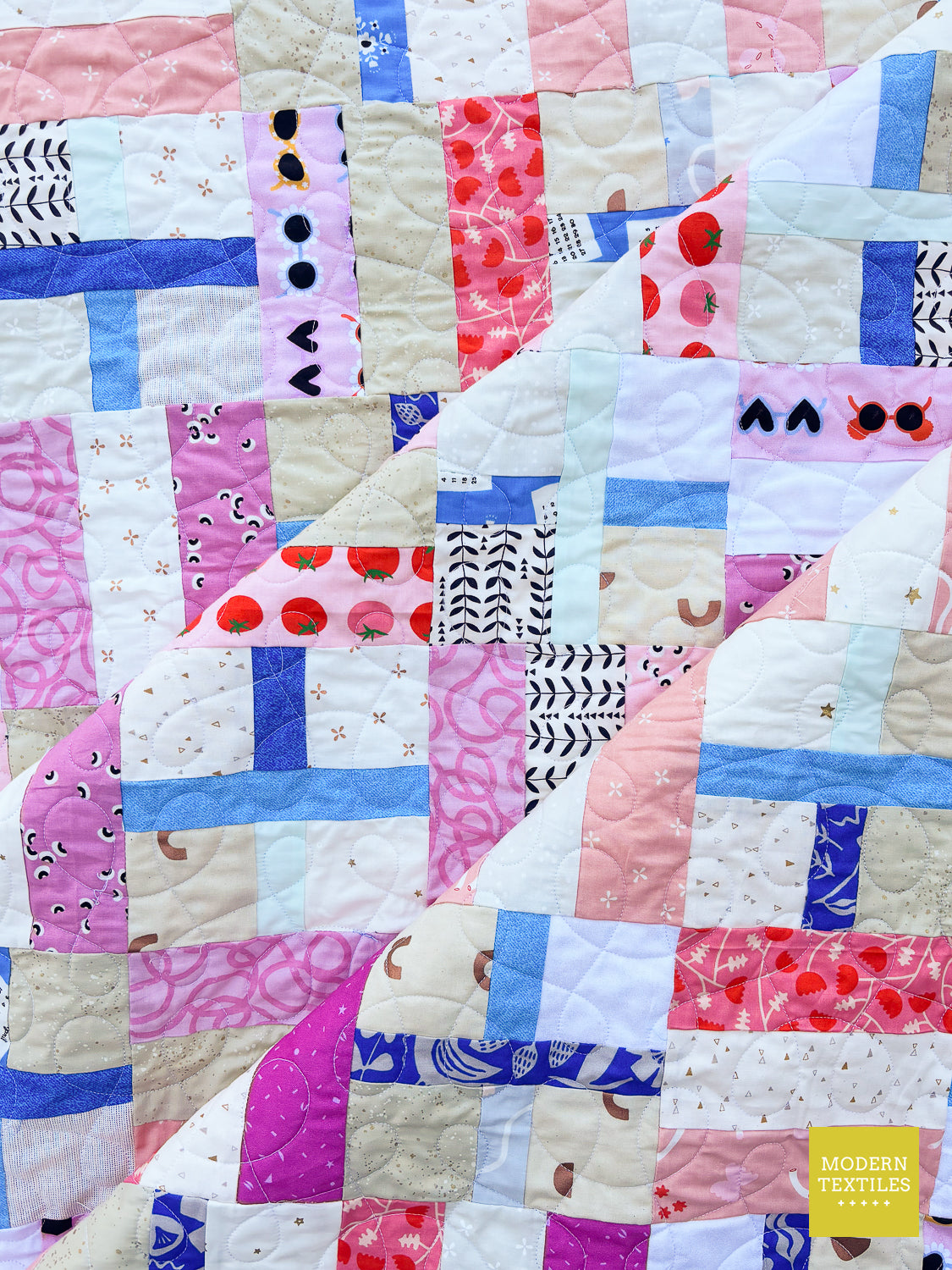 A close-up of a patchwork quilt featuring various fabric panels in pink, blue, beige, and white, including patterns of strawberries, dots, stripes, and floral prints. A yellow 'MODERN TEXTILES' logo is visible in the bottom right corner.