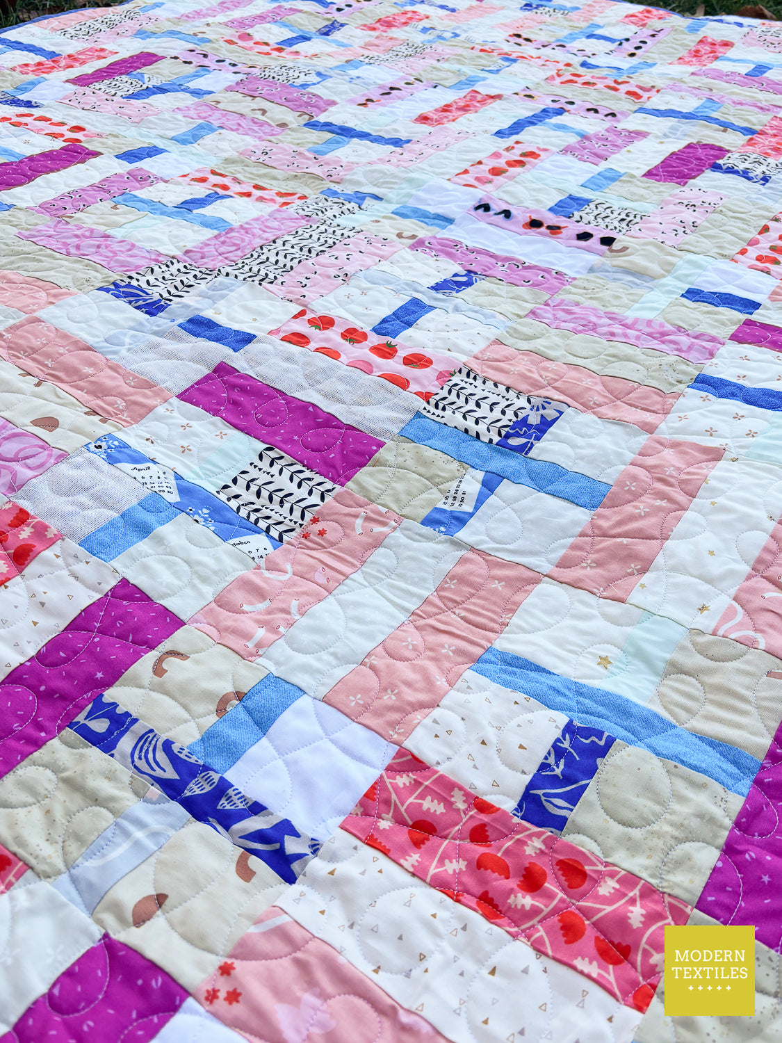 A colorful patchwork quilt laid flat, featuring a variety of fabric patterns and colors including pink, blue, purple, and white. The quilt has a quilted texture and is photographed outdoors with a yellow 'Modern Textiles' logo in the bottom right corner.