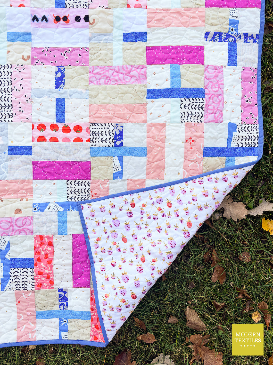 A colorful patchwork quilt laid outdoors on grass, with one corner folded back to show the printed fabric backing. The quilt features a variety of pink, blue, white, and beige patterned fabric squares with a blue border.