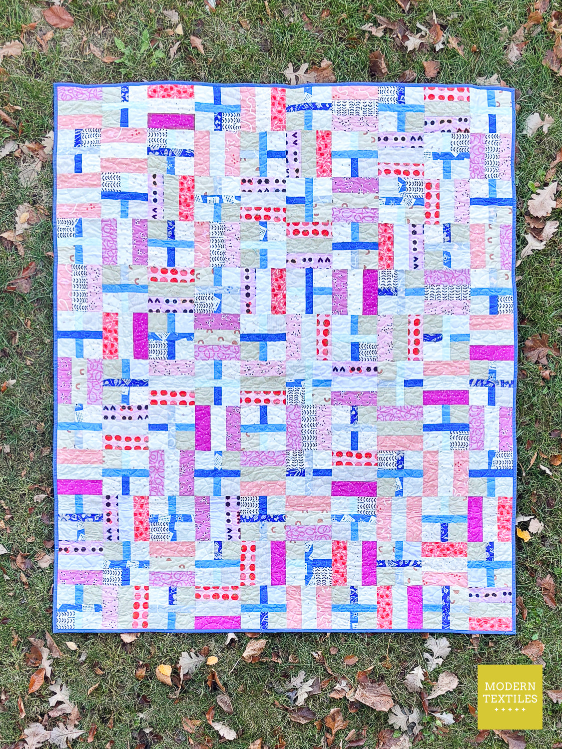 A colorful patchwork quilt laid flat on grass with scattered fallen leaves. The quilt features a geometric block pattern in shades of blue, pink, purple, and white.