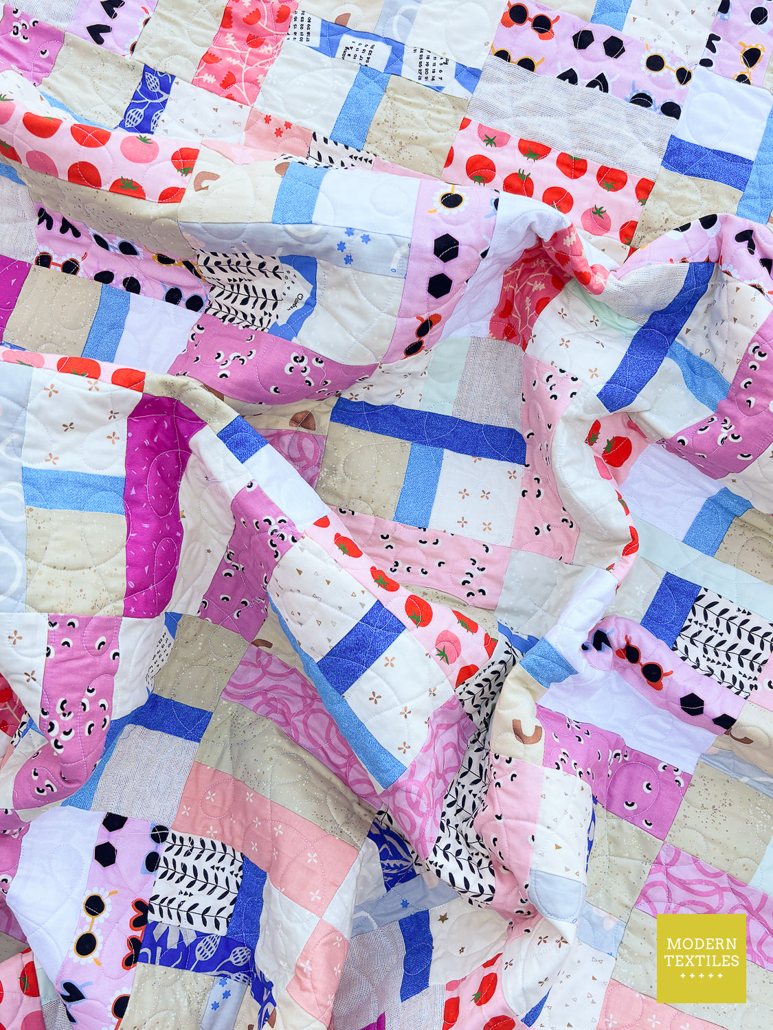 A close-up of a colorful patchwork quilt with various fabric prints including pink, blue, red, black, and beige, featuring a small yellow 'Modern Textiles' logo in the bottom right corner.