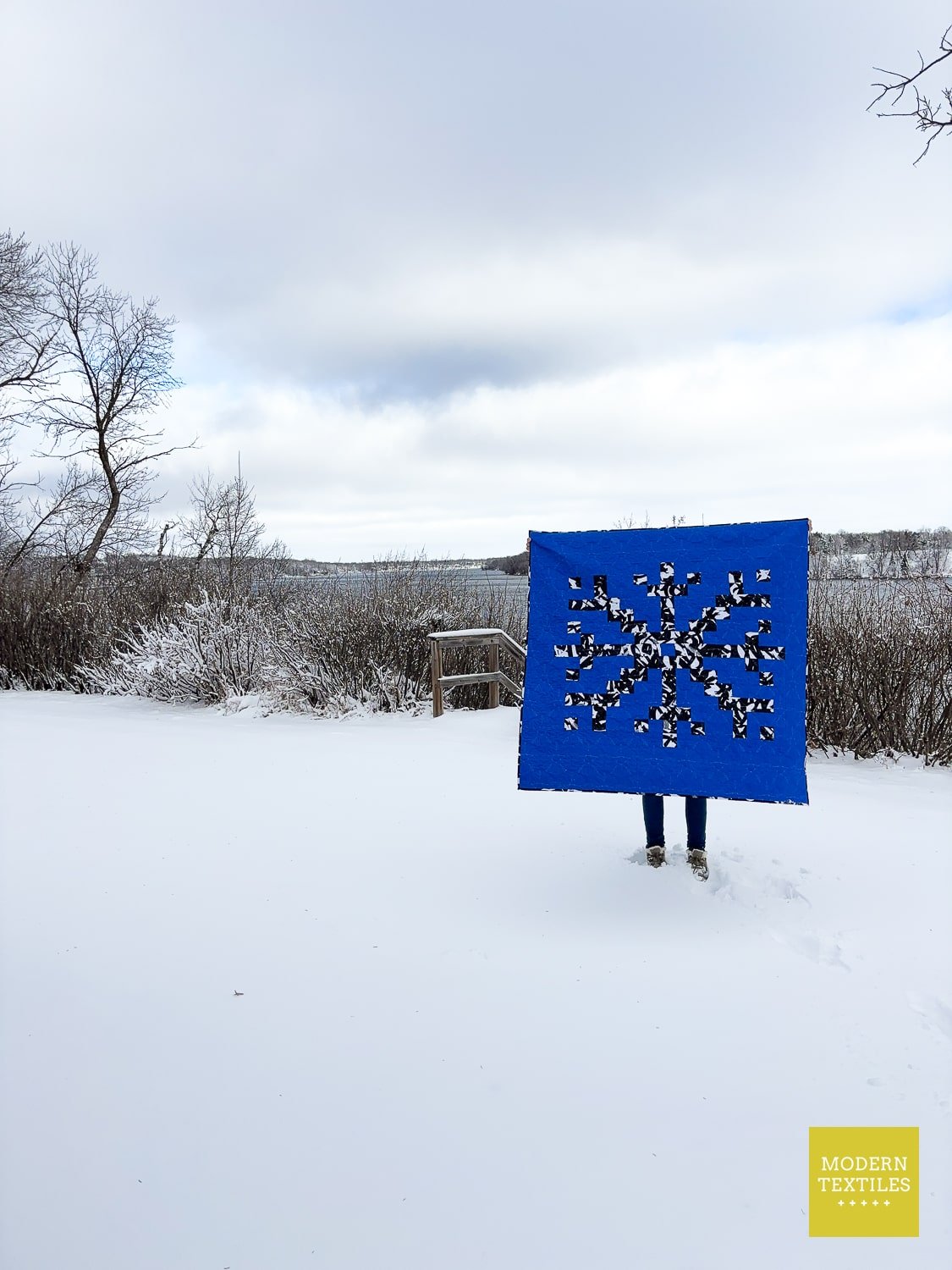 Person holding a snowflake quilt with snowflake design in a snowy landscape.