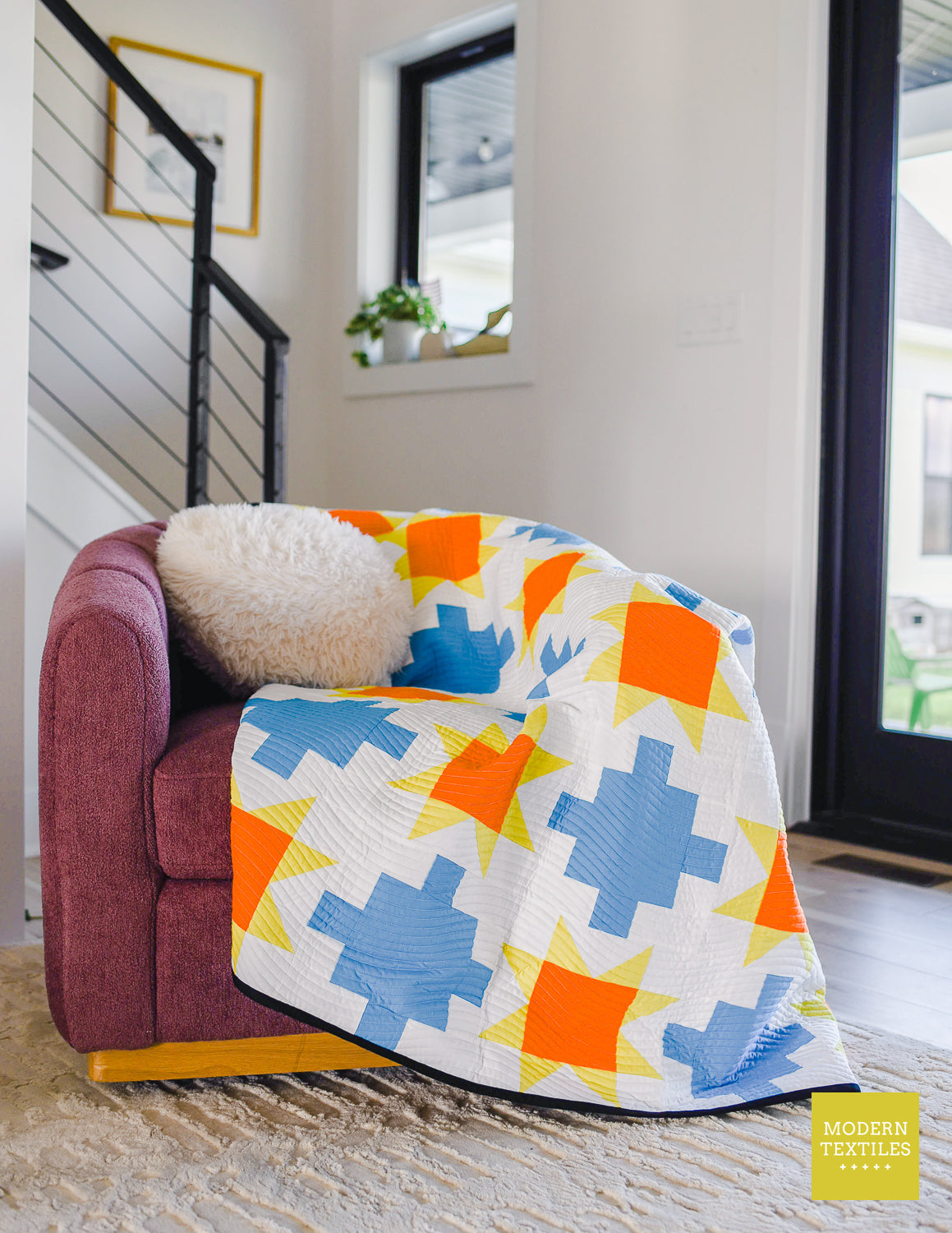 Close-up of blue patchwork quilt with sawtooth stars in orange and yellow-green with white background fabric and black binding fabric draped across maroon colored chair with white fluffy pillow on top of quilt.