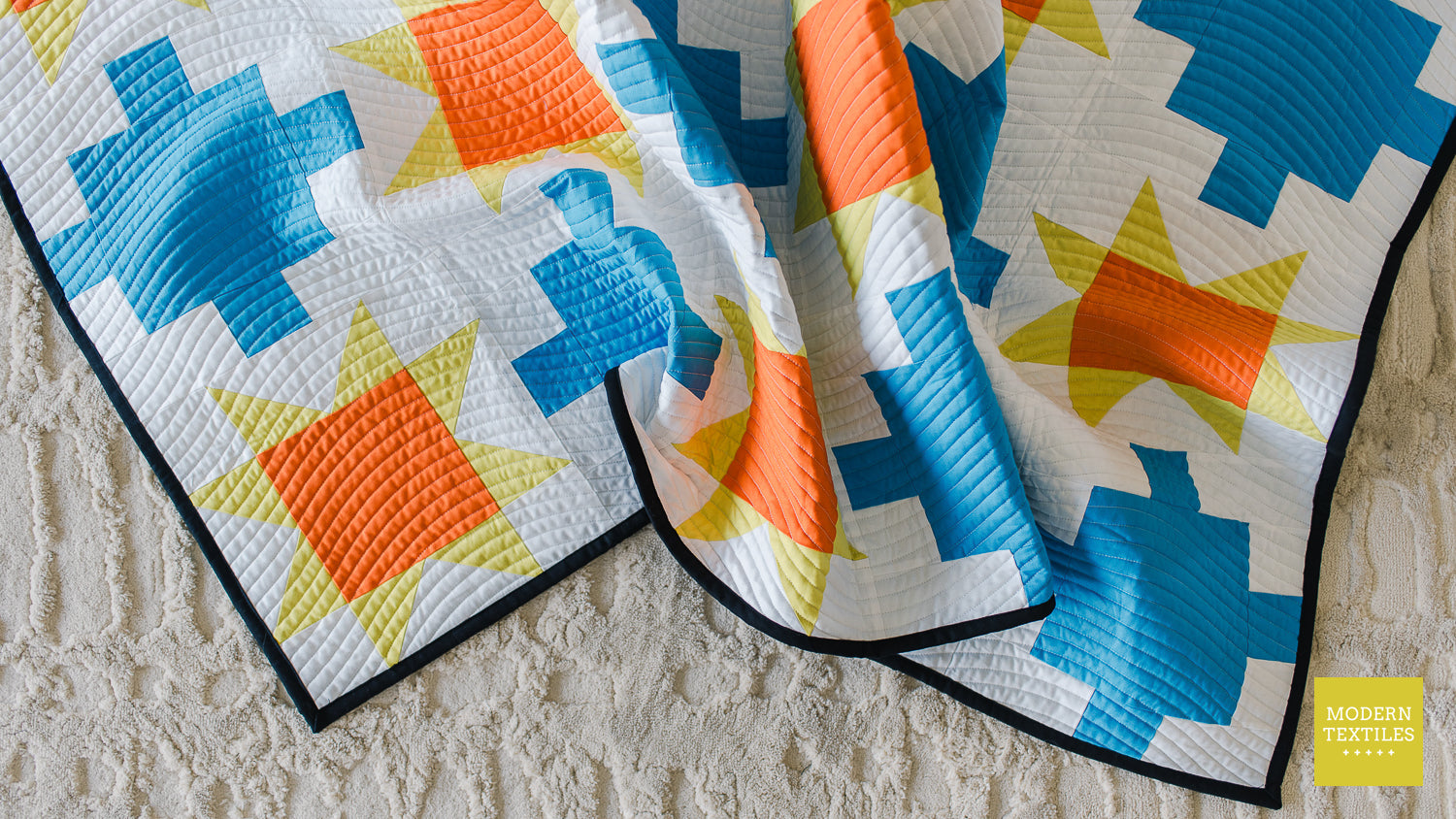 Close-up of blue patchwork quilt with sawtooth stars in orange and yellow-green with white background fabric and black binding fabric crinkled on top of a neutral textured rug.
