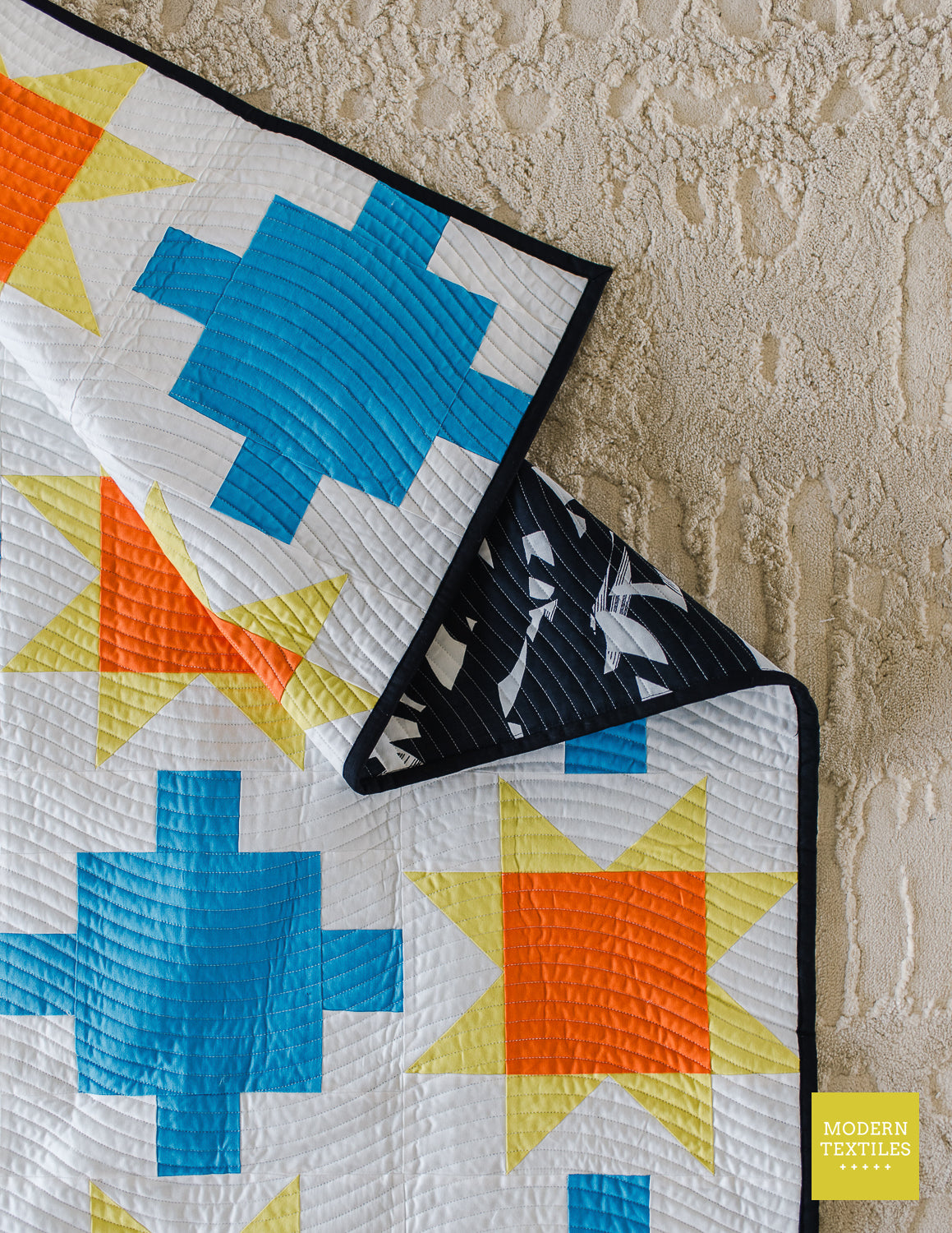 Close-up of blue patchwork quilt with sawtooth stars in orange and yellow-green with white background fabric and black binding fabric with black and white brush stroke backing fabric showing to the front. Quilt on top of neutral textured rug.