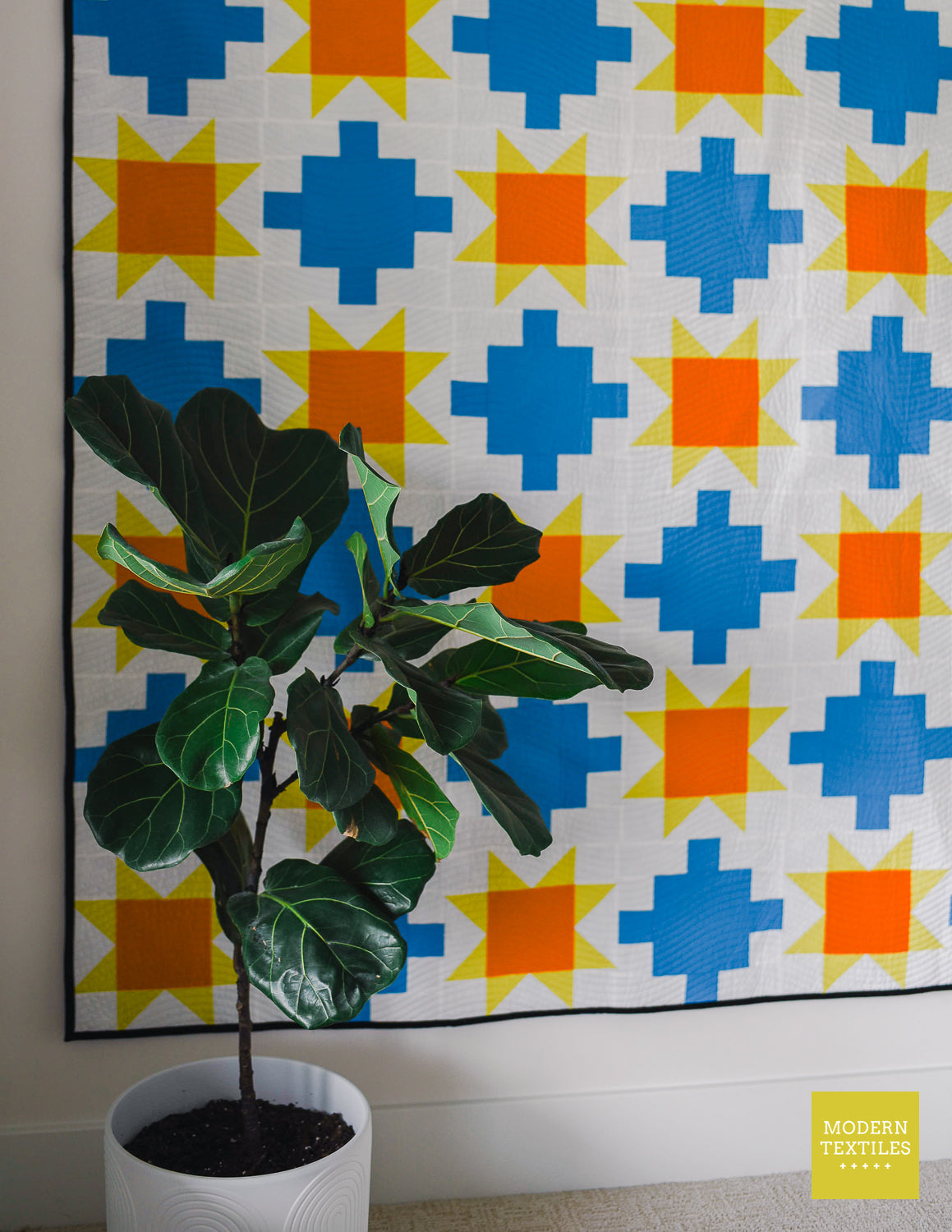 Potted fig plant in front of a sawtooth star and patchwork quilt on white wall.