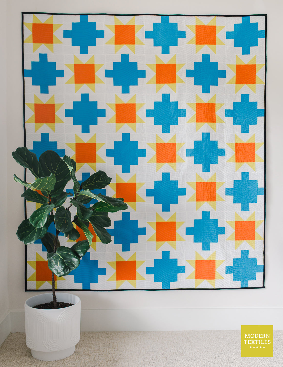 Blue patchwork quilt with sawtooth stars in orange and yellow-green geometric-patterned quilt on a white wall with a potted fig plant in the foreground.