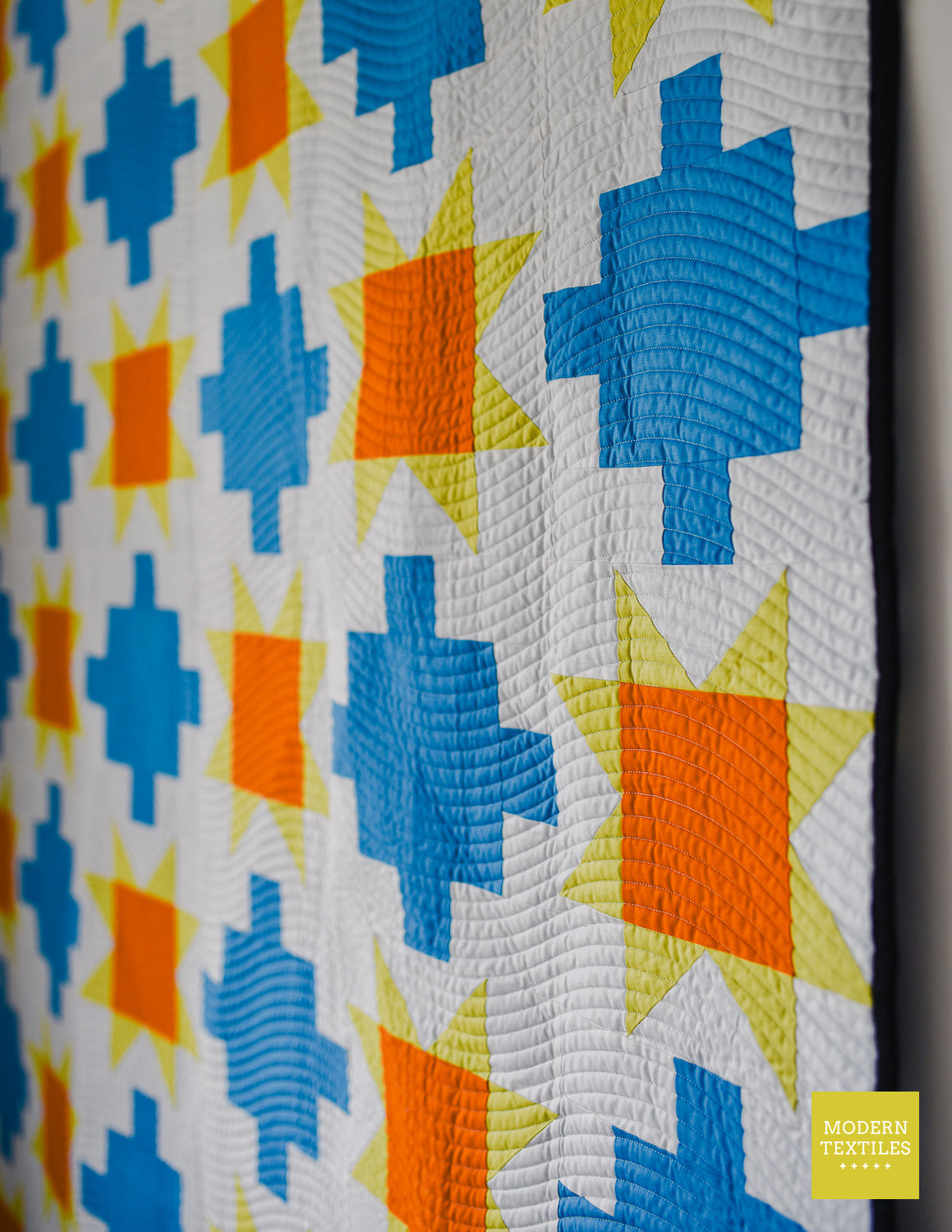 Blue patchwork quilt with sawtooth stars in orange and yellow-green geometric-patterned quilt on a white wall showcasing wavy lines quilting.