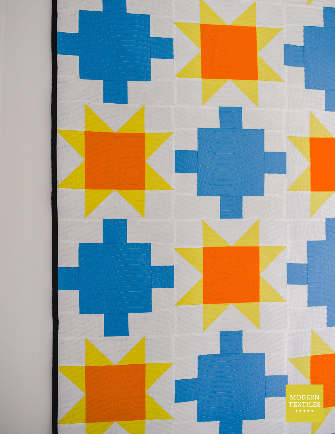 Close-up of blue patchwork quilt with sawtooth stars in orange and yellow-green with white background fabric and black binding quilt on a white wall.