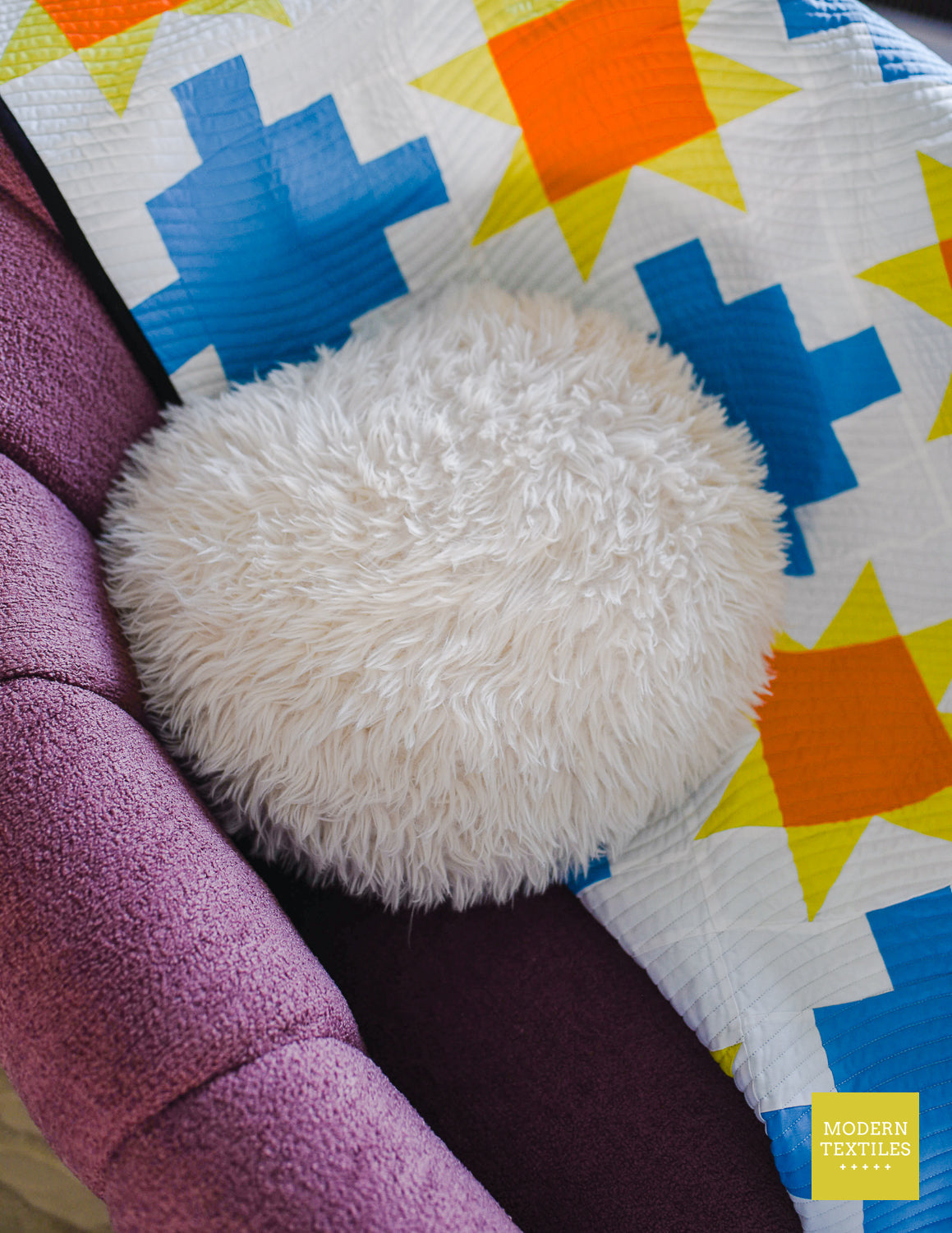 Blue patchwork quilt with sawtooth stars in orange and yellow-green with white background fabric and black binding quilt on a plum colored chair with a fluffy white round pillow on top of the quilt.