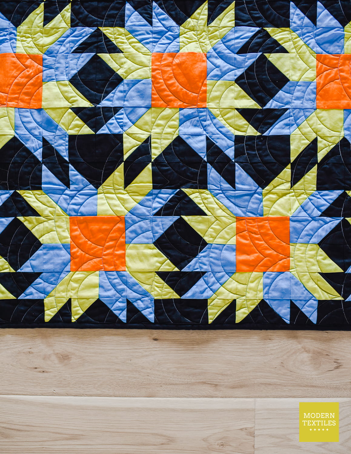 A close-up of a quilt texture featuring a colorful geometric starburst pattern with black, blue, lime green, and orange blocks, laid flat on a light wood floor. Highlighting the off-center block placement.