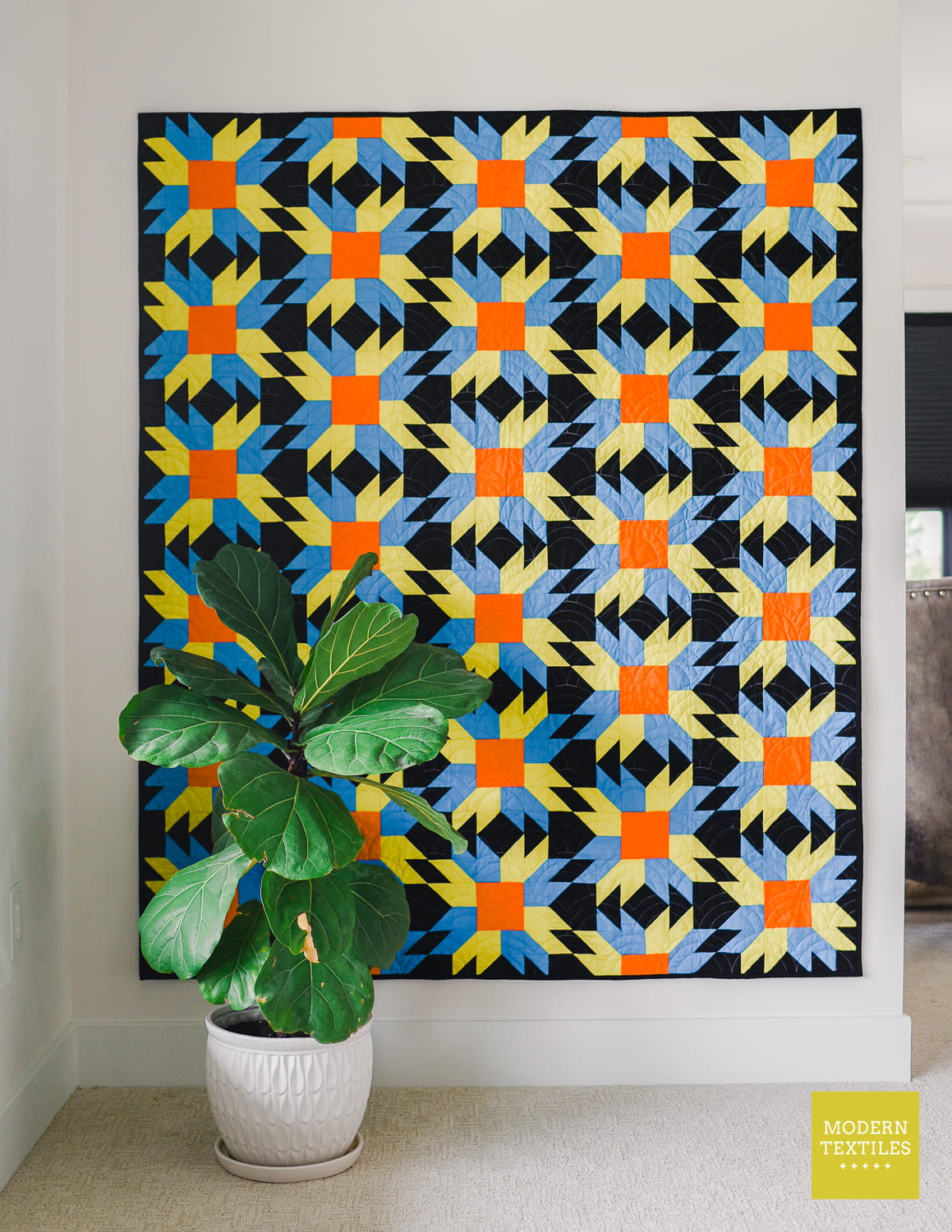 A throw size quilt with a bold geometric starburst pattern in black, green, blue, and orange, displayed in a room corner with a potted green plant in front and a visible sofa on the right background. 