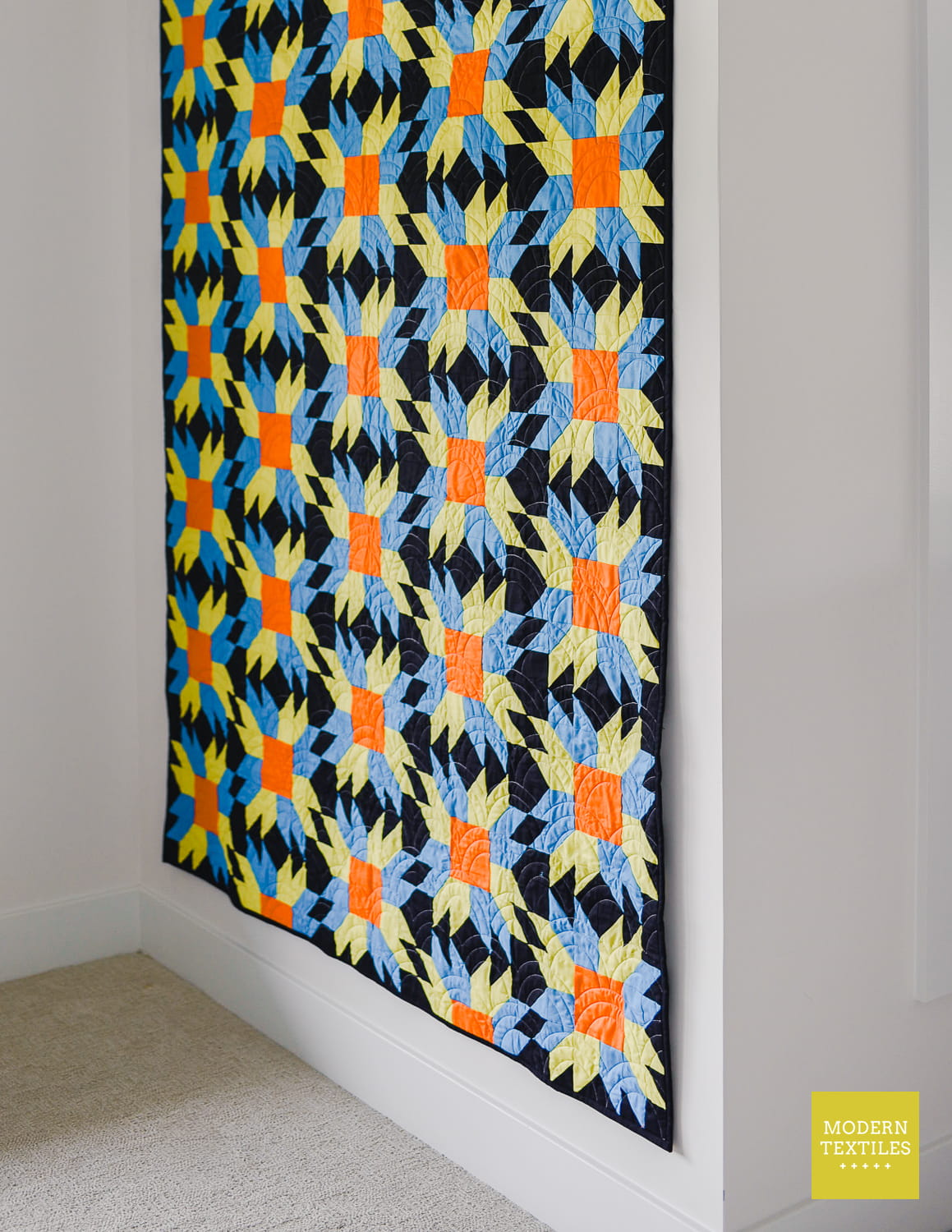 A colorful throw size quilt used as wall hanging with a geometric Sunfire pattern in black, blue, green, and orange, displayed on a white wall in a room corner.